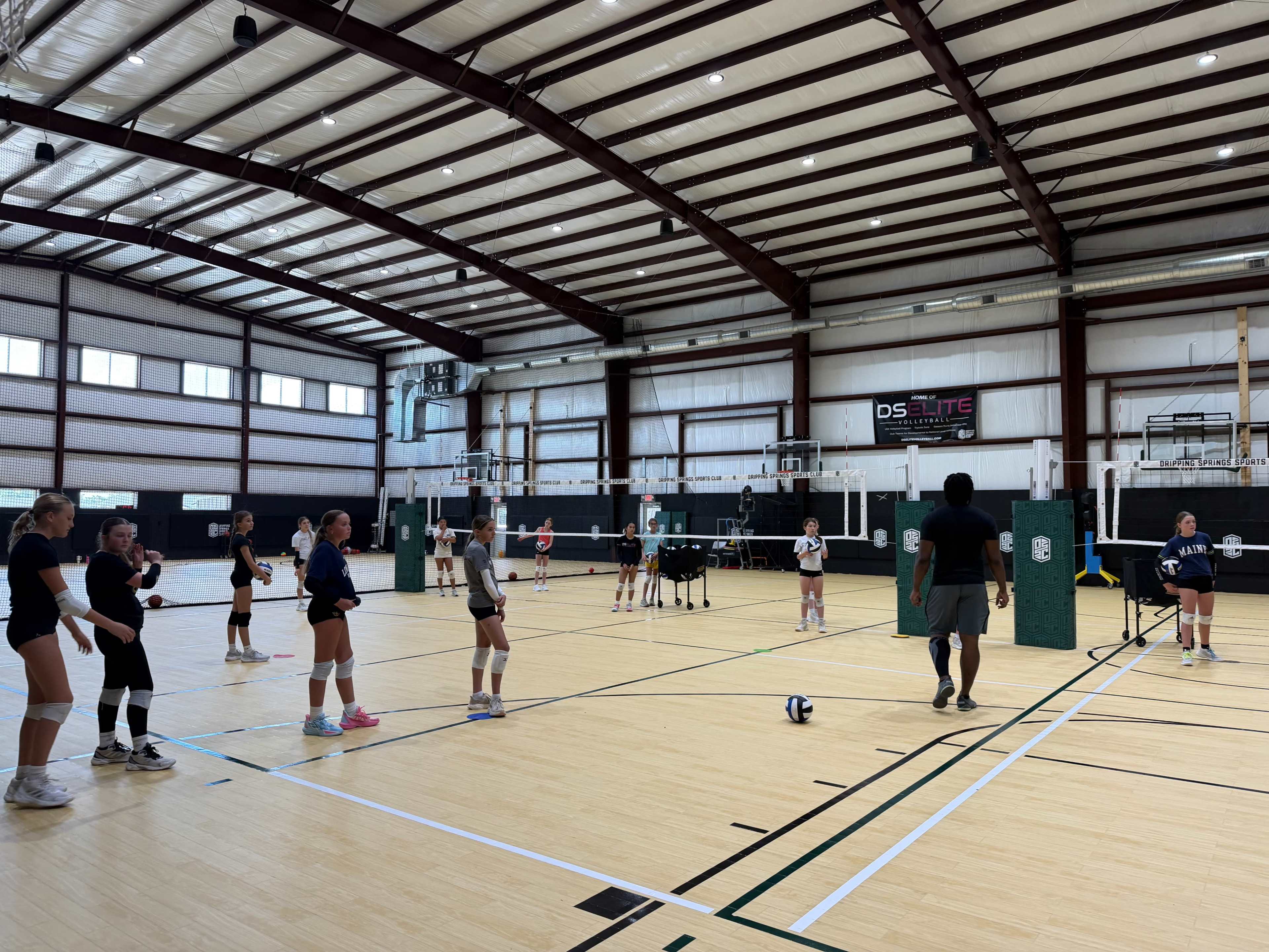 A volleyball practice session takes place in a large indoor gymnasium, featuring several players positioning themselves around a central court with nets and volleyballs.