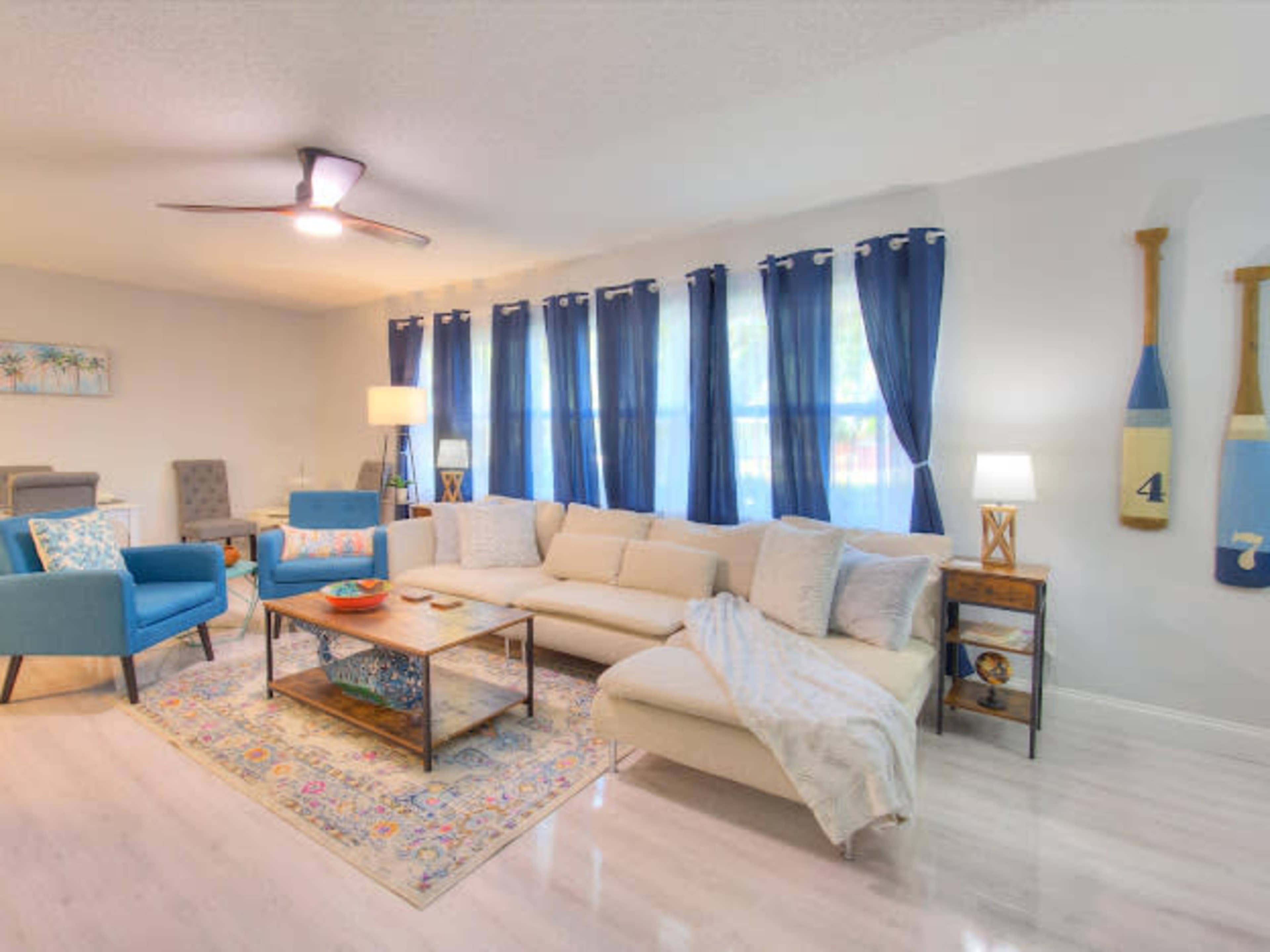 The image shows a bright living room with a sectional couch, blue armchairs, a coffee table, and large windows covered with blue curtains.