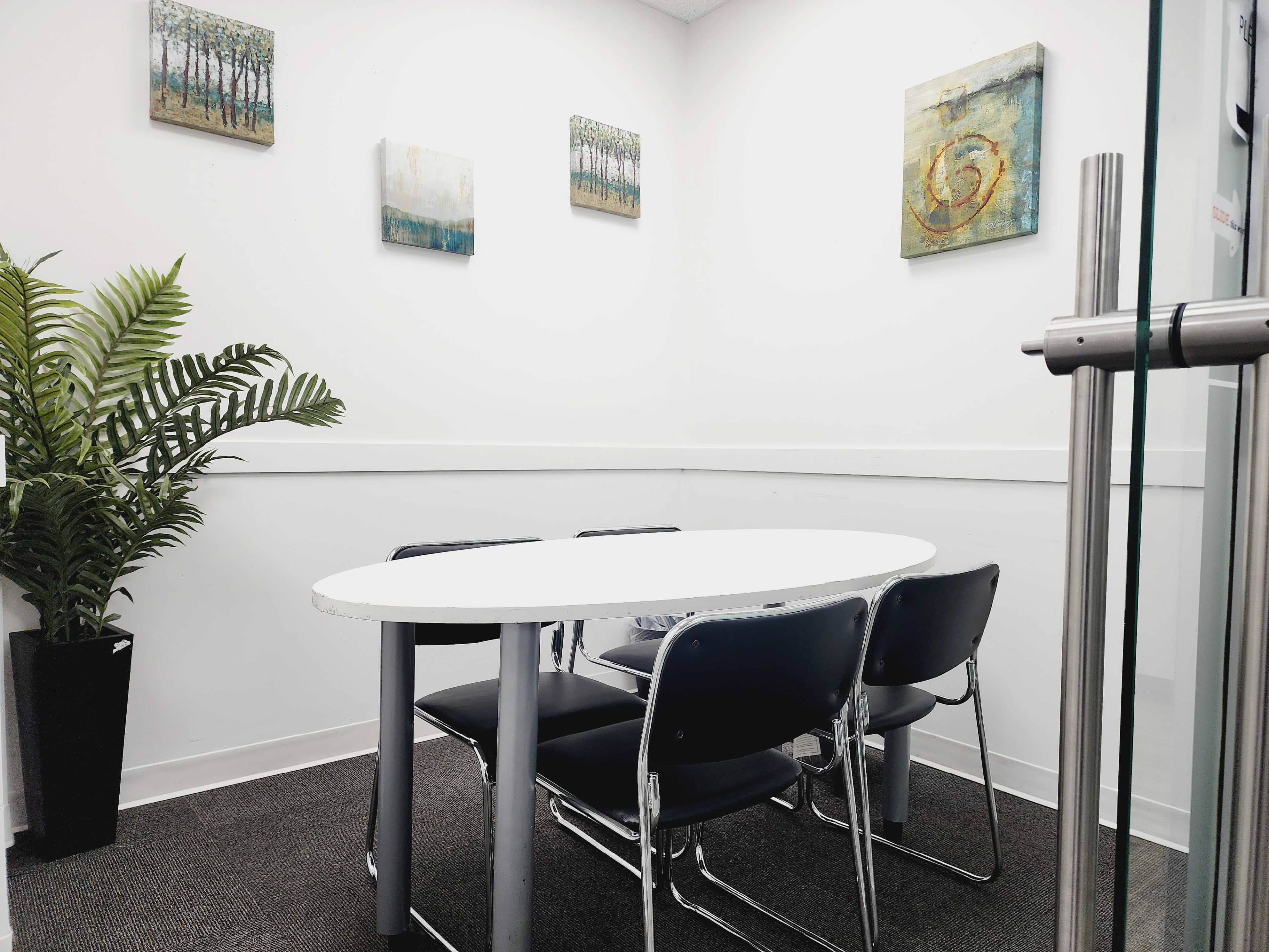 A small meeting room features a round table surrounded by three chairs, with a potted plant in one corner and artwork on the walls.