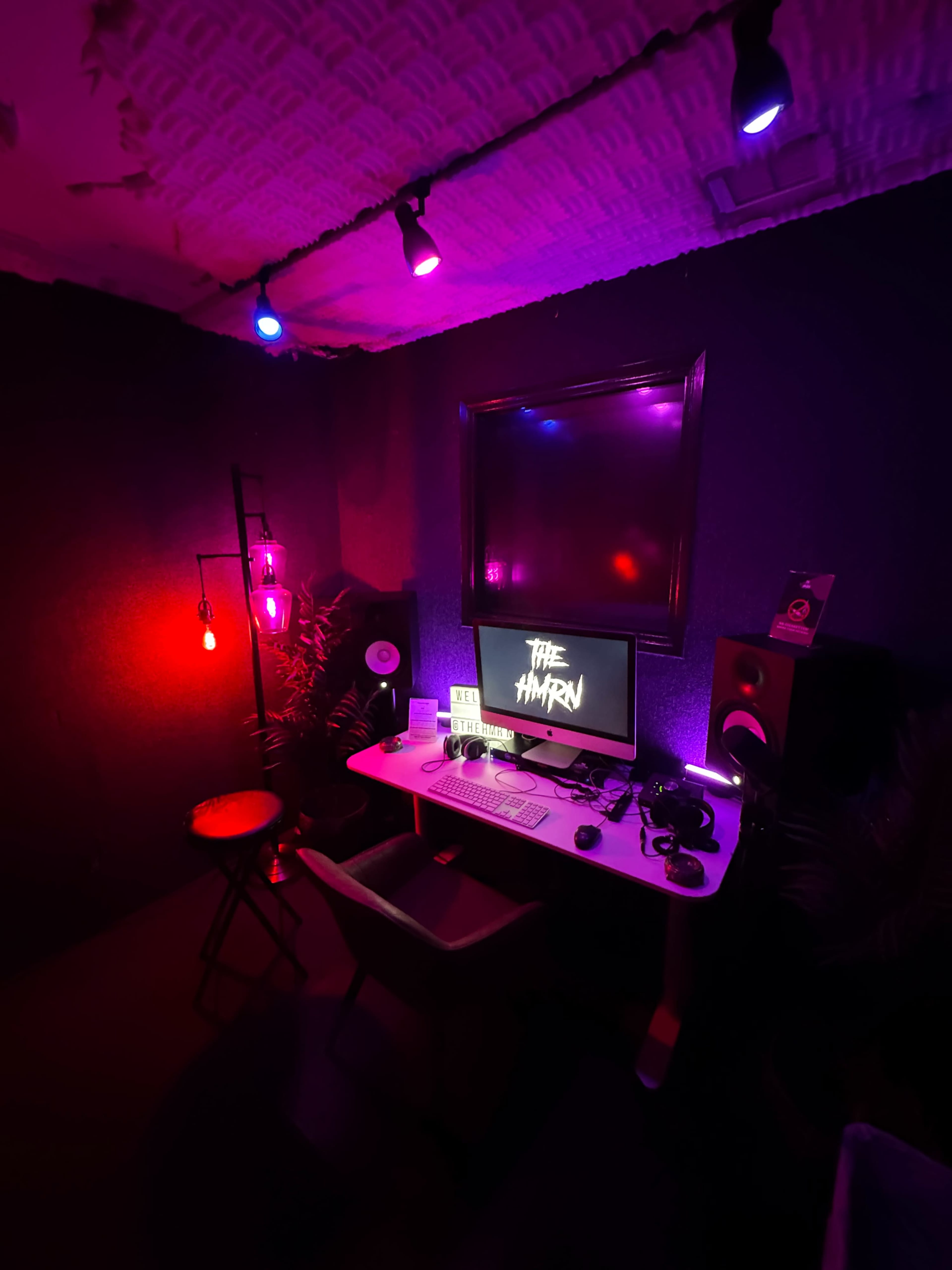 A dimly lit room features a desk with a computer monitor, speakers, and a chair, illuminated by colorful lighting from overhead and a hanging lamp.