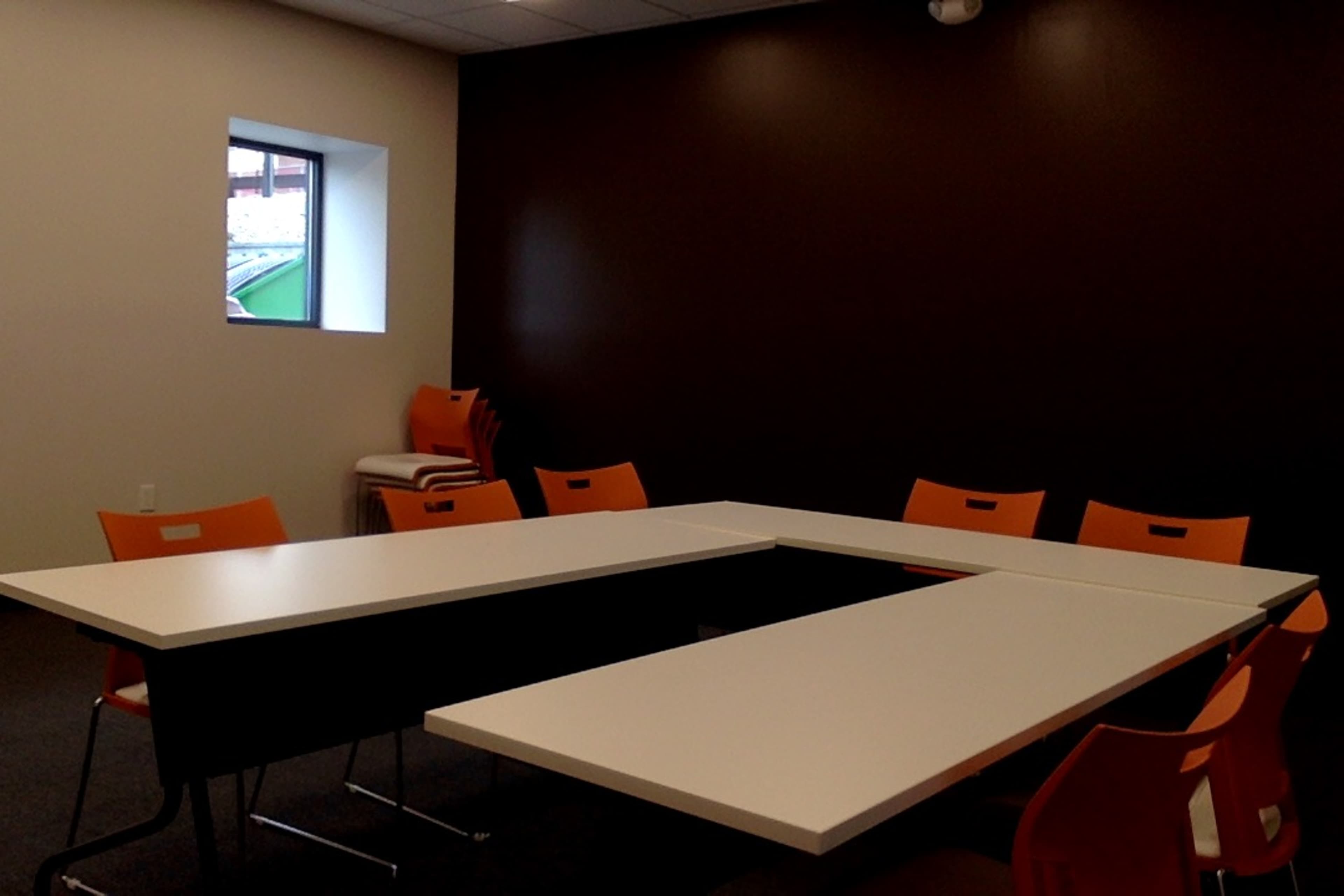 A conference room features two white tables arranged in a T shape, surrounded by orange chairs, with a window showing a view outside.