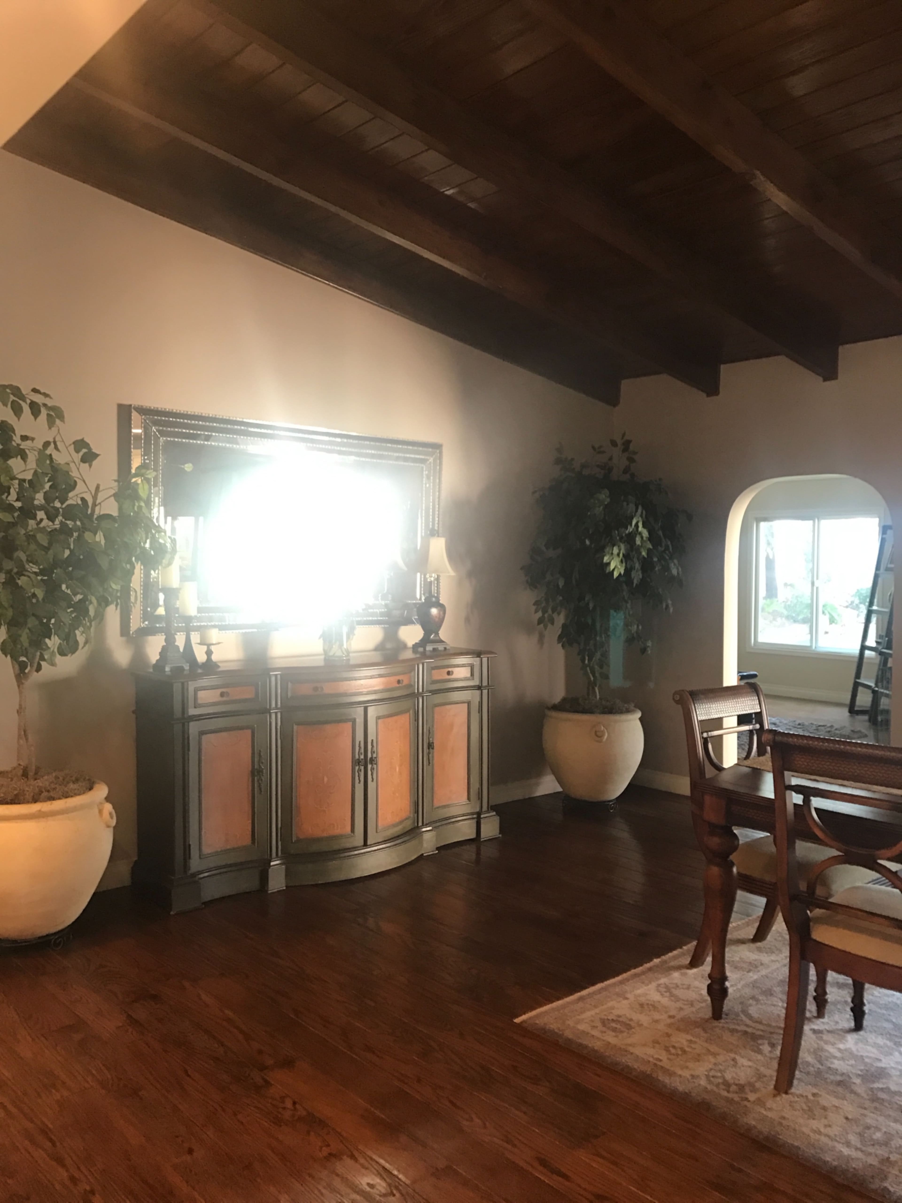 A wooden sideboard stands against a wall with a large mirror above it, flanked by potted plants, while a dining table sits nearby in a room with wooden beams on the ceiling.