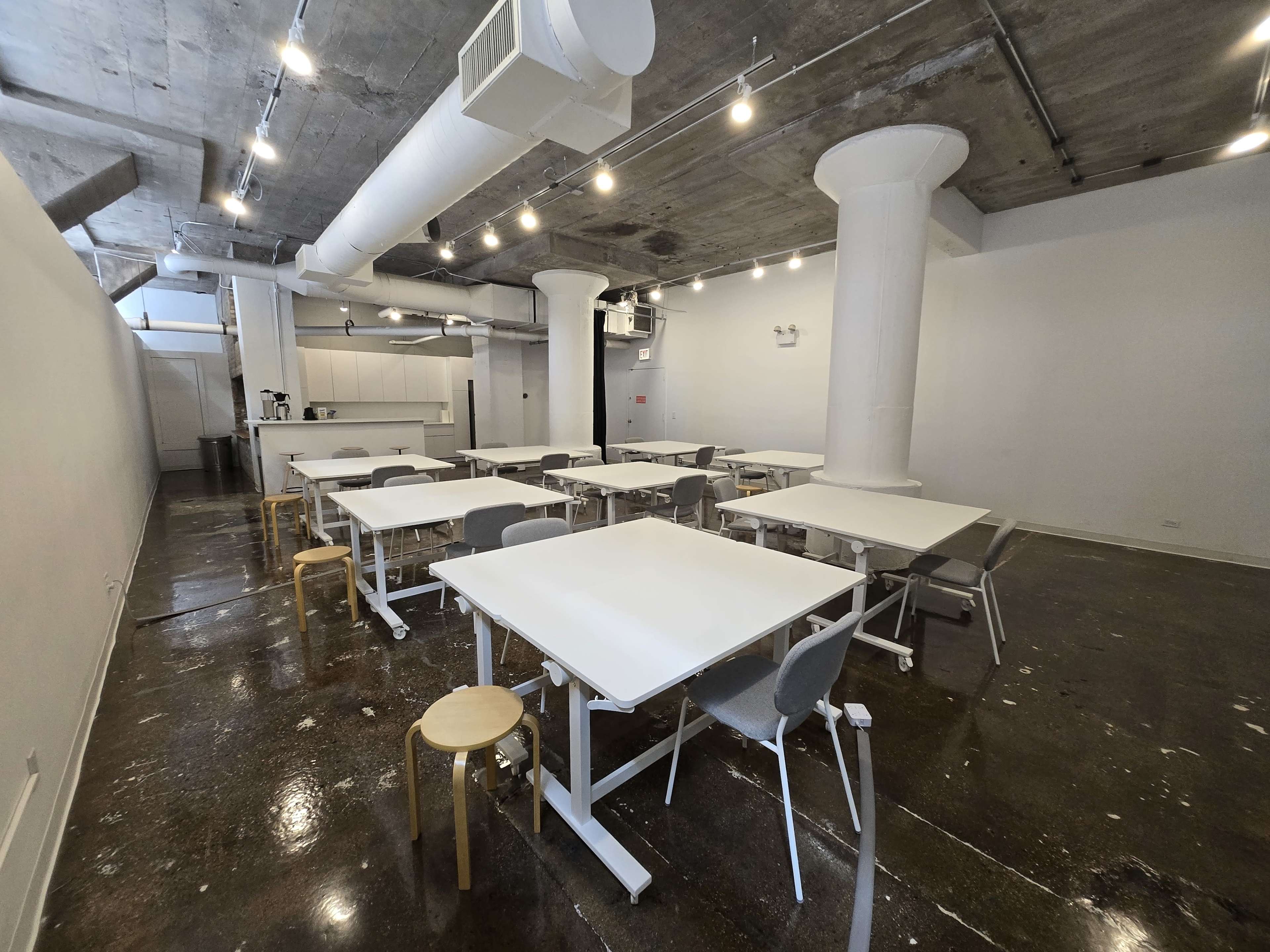 The image shows a spacious, modern room with multiple tables and chairs arranged in a grid pattern, concrete walls, and an industrial ceiling with exposed pipes.