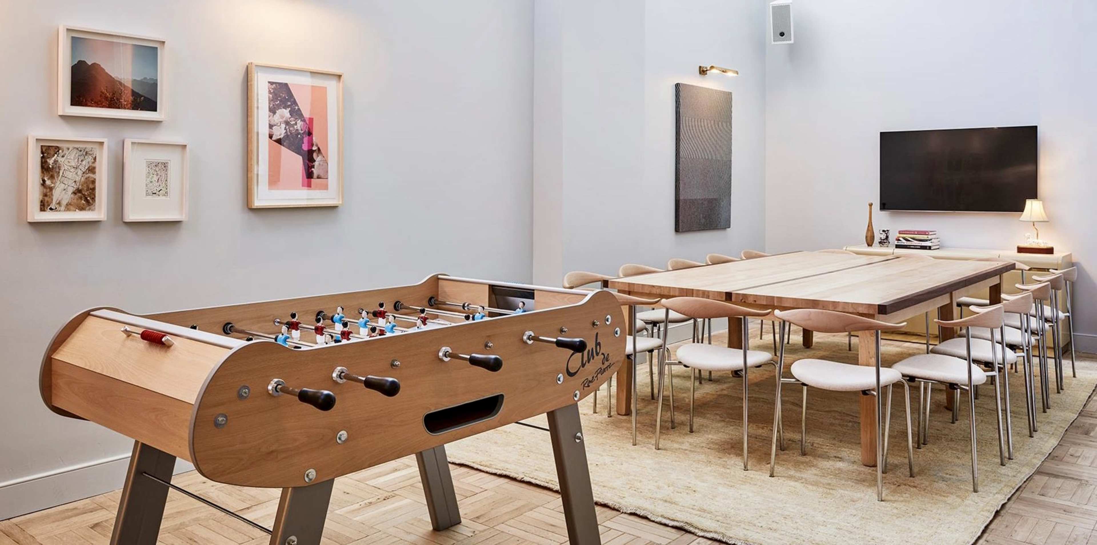 The image shows a spacious room with a foosball table in the foreground and a long dining table surrounded by chairs in the background.