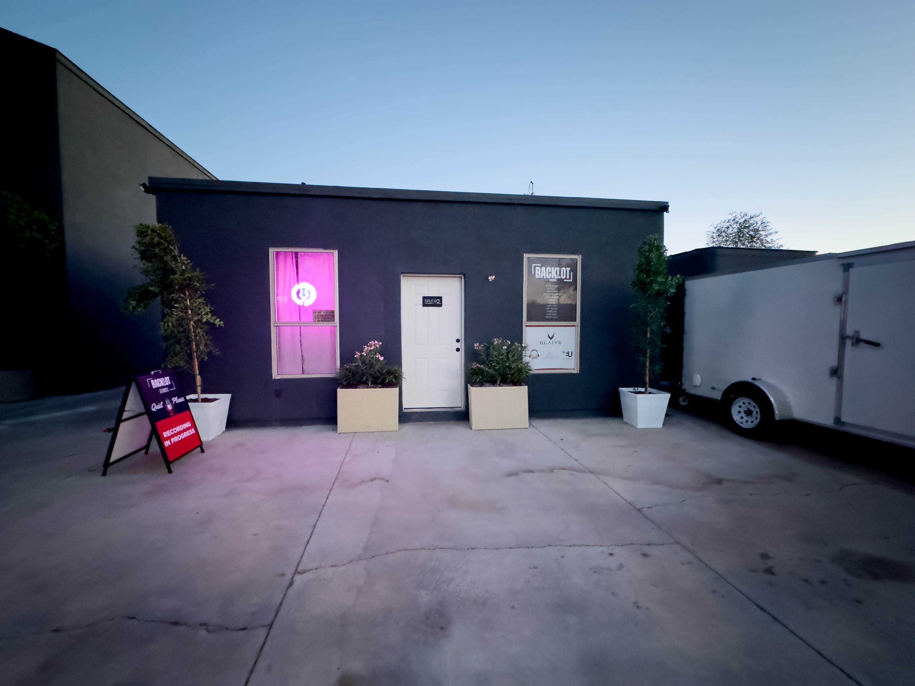 The image shows a small gray building with a white door, flanked by potted plants, and illuminated by purple lighting from a window.