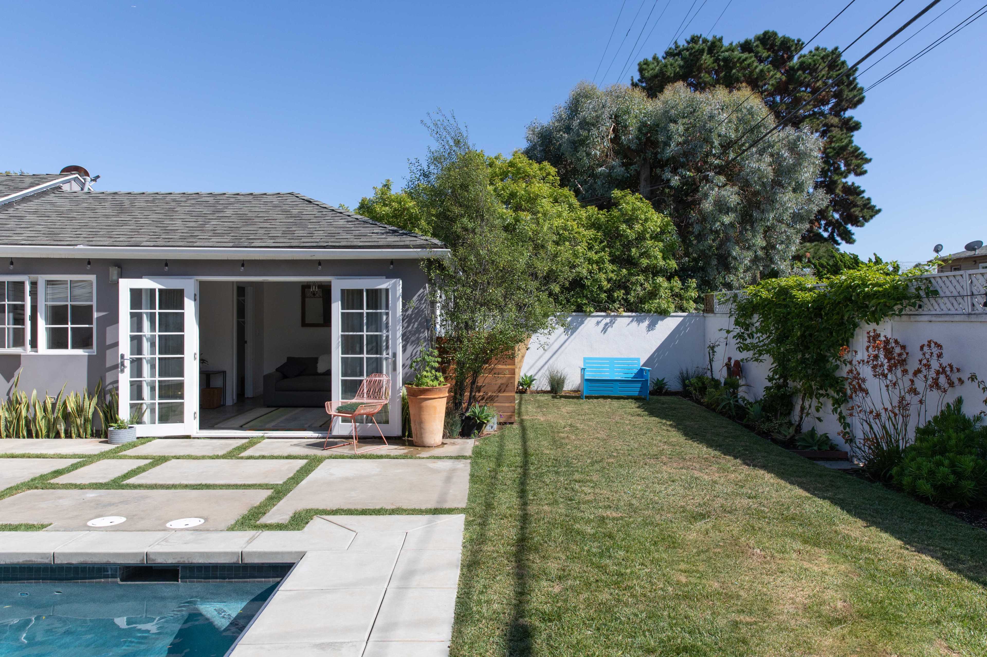 The image shows a backyard with a pool, a patio area featuring a seating arrangement, and a grassy lawn bordered by plants and shrubs.