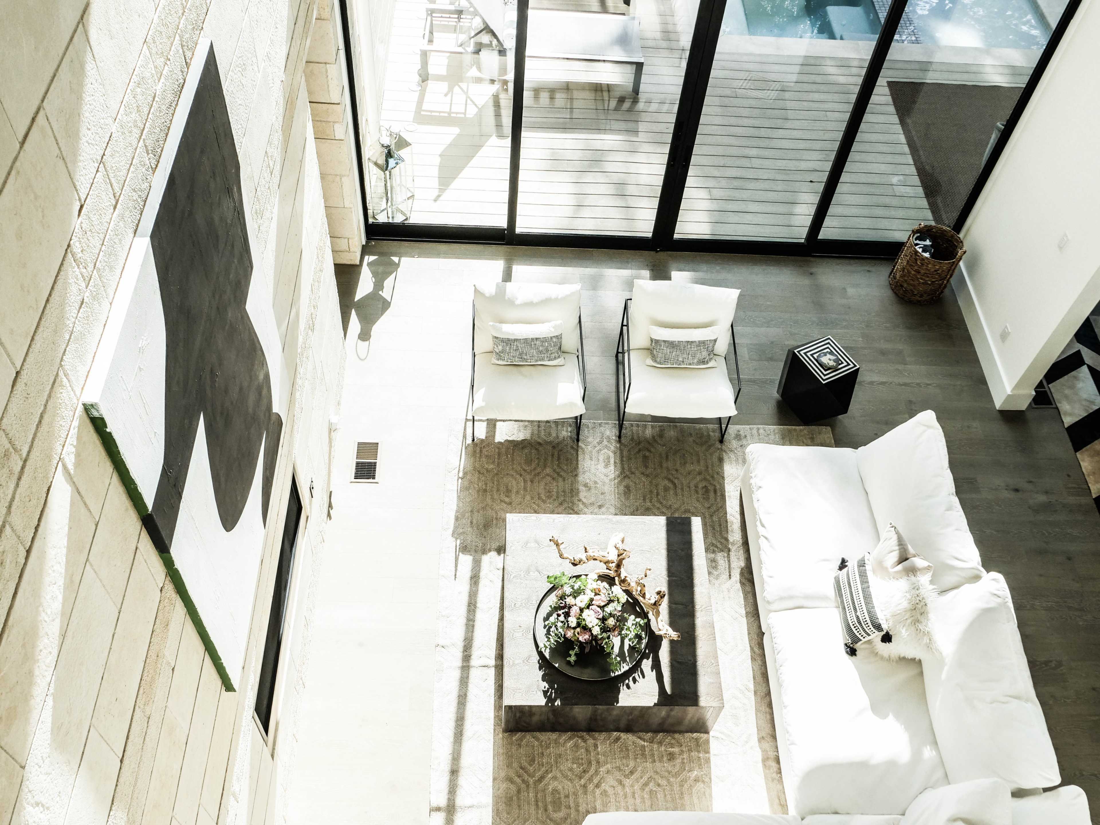 A spacious living room with a large white sofa, two chairs, a coffee table with a floral arrangement, and a large artwork on the wall, all surrounded by large windows that offer views of an outdoor deck.