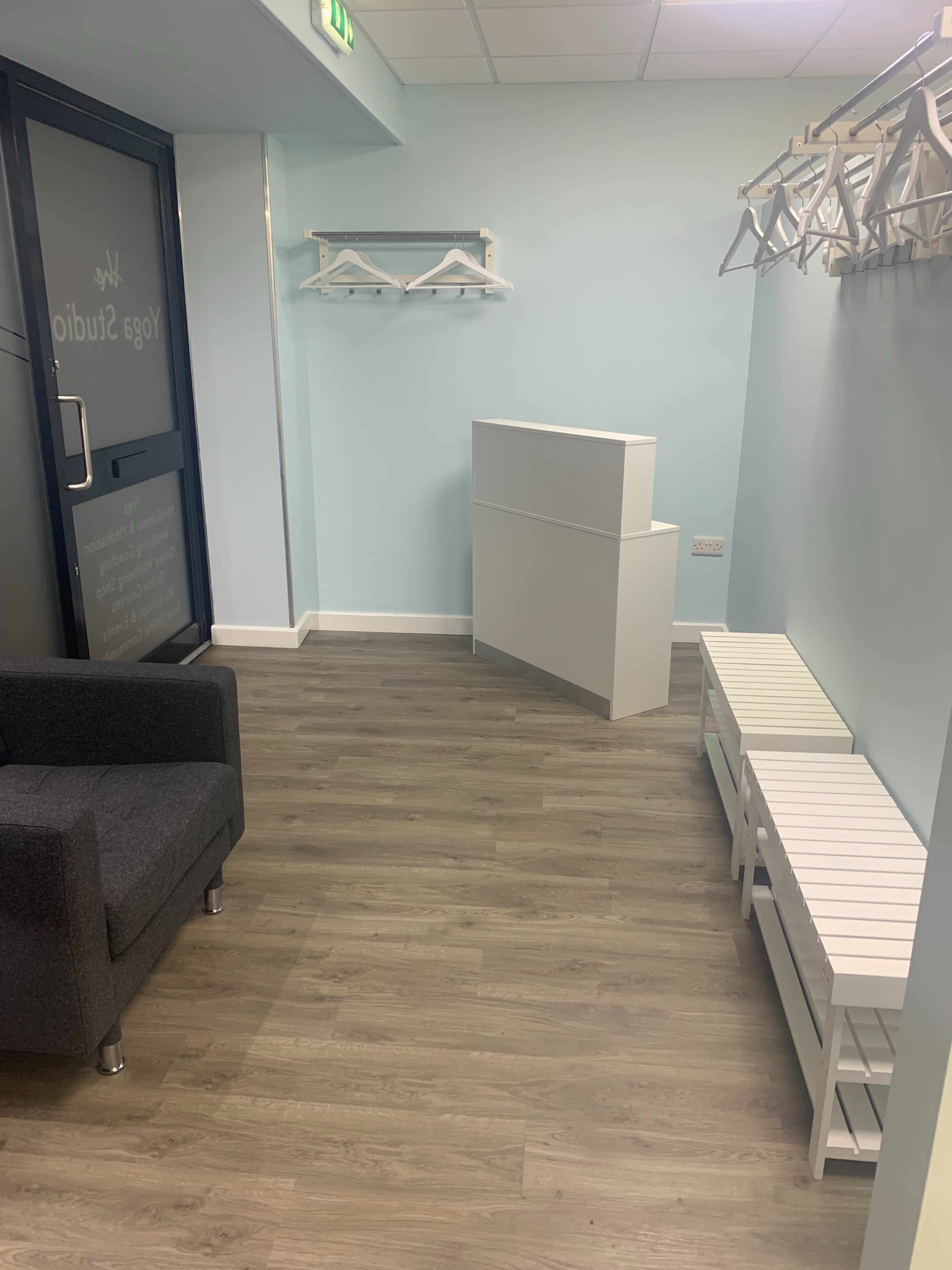 A minimalistic room featuring a gray armchair, a white reception desk, and several white coat hangers on the wall.