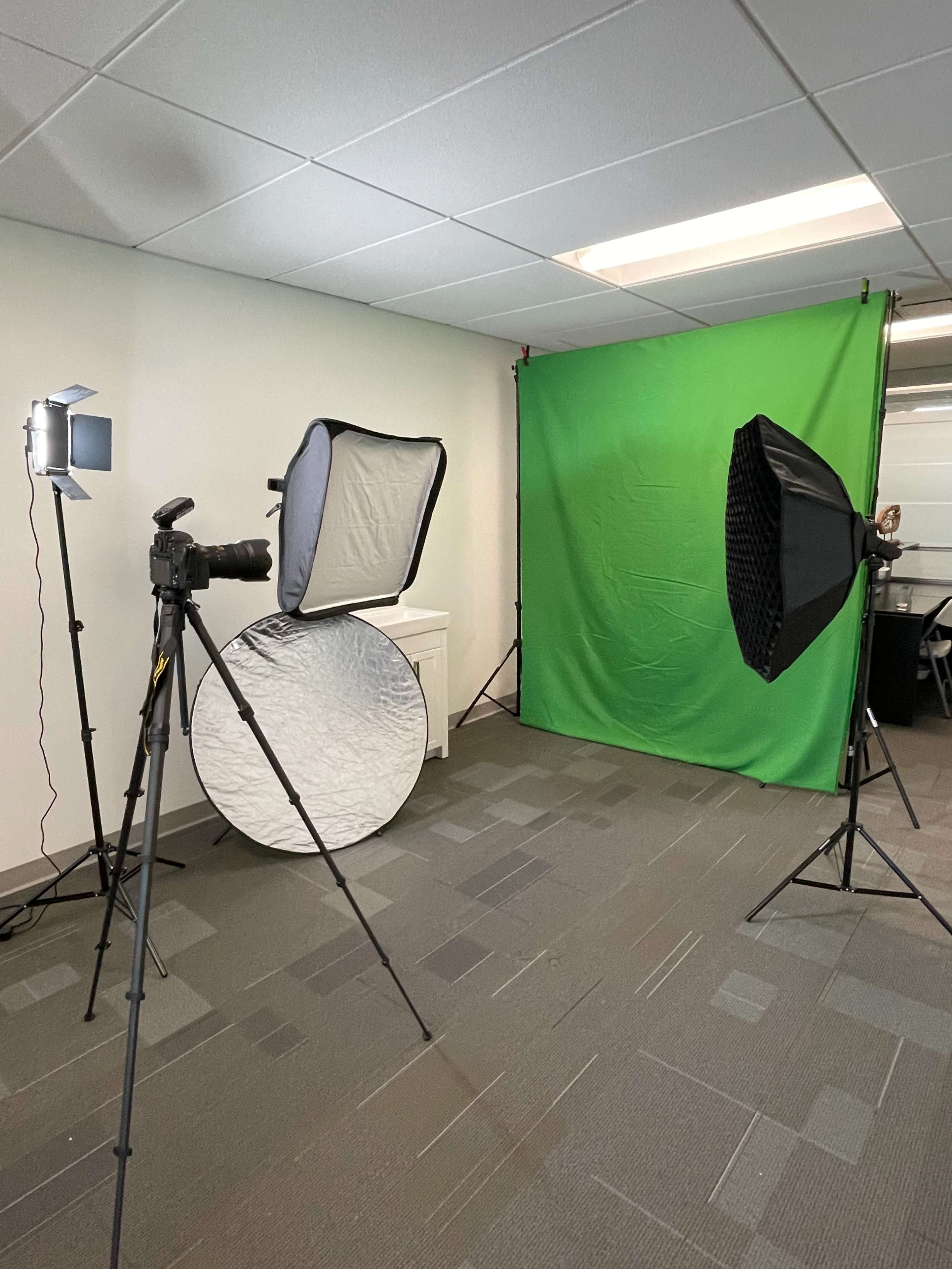 A photography setup featuring a green screen, softbox lights, a camera on a tripod, and a reflective disc in a room with gray carpeting.