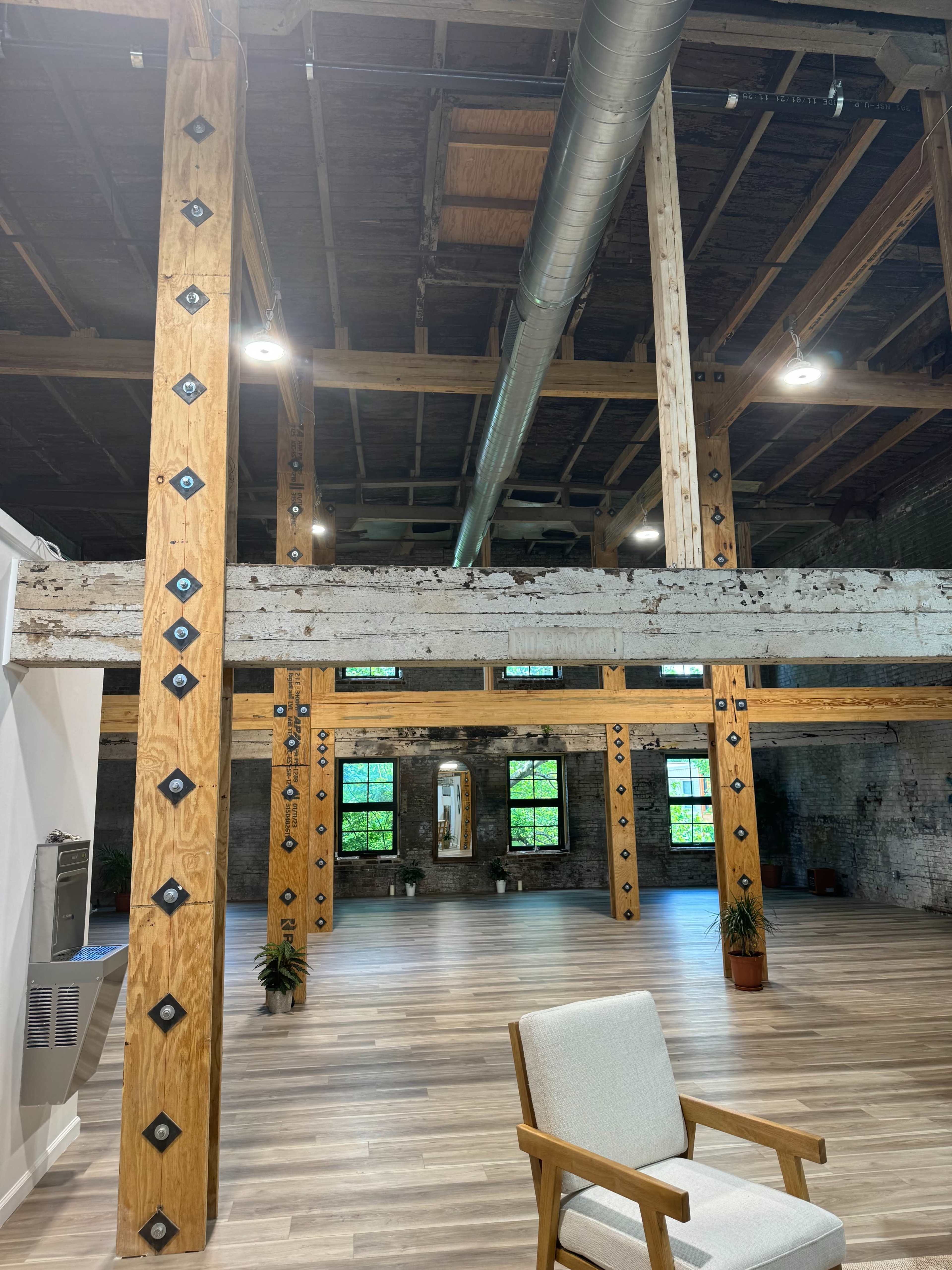 An industrial-style interior space with exposed wooden beams, metal accents, and large windows overlooking a wooden floor.