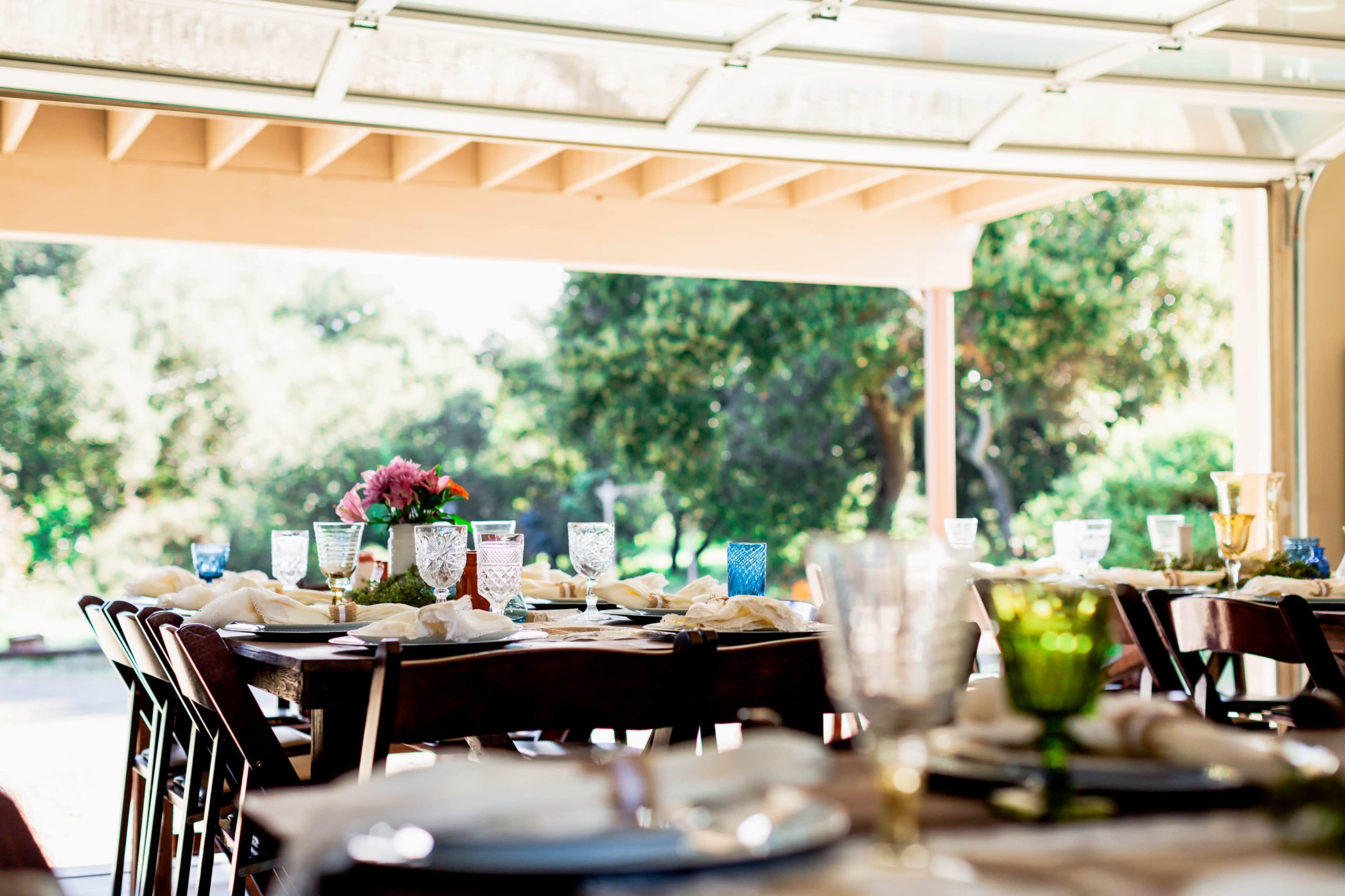 A dining area is set with plates and colorful glassware, overlooking a garden through large open doors.