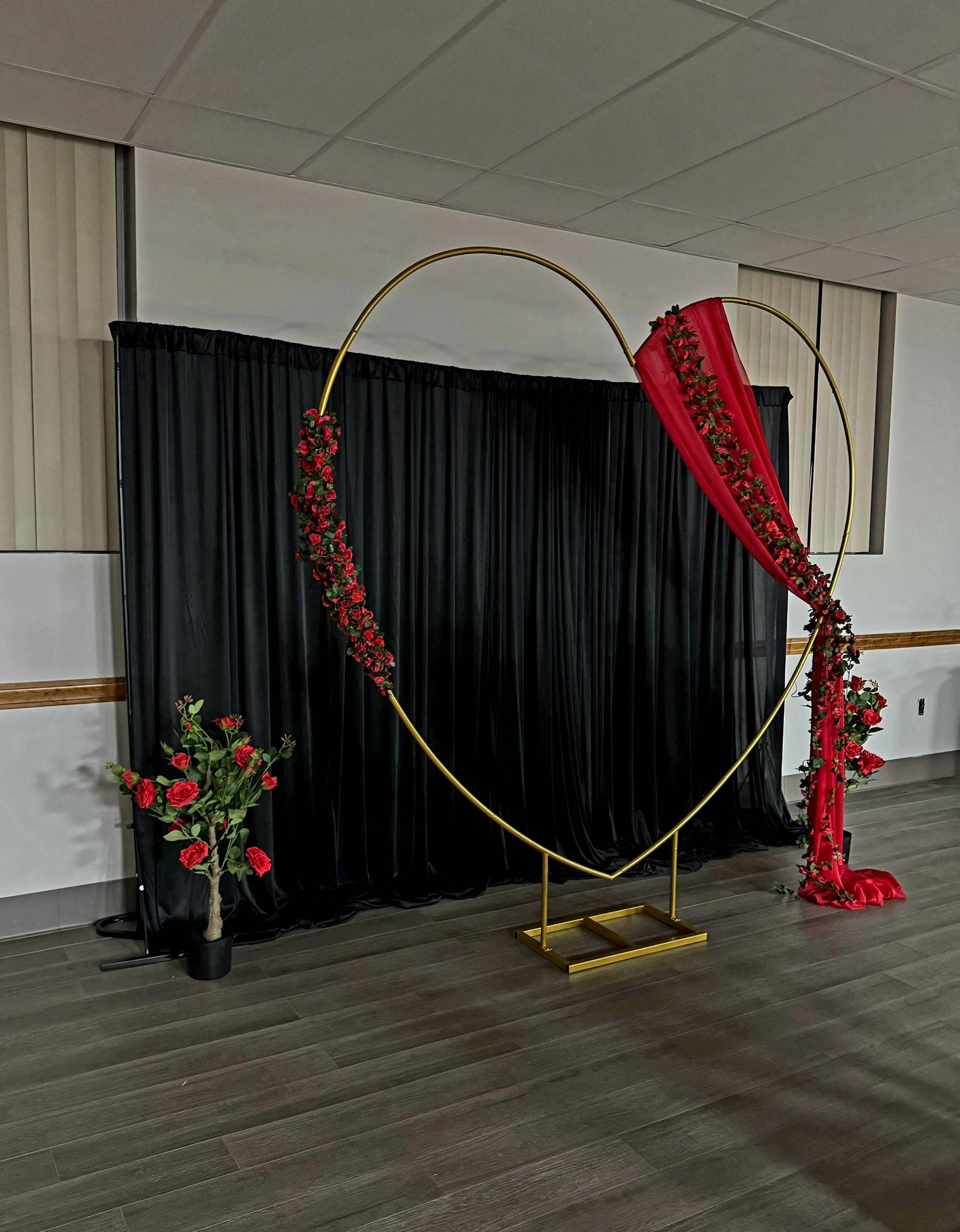 A large heart-shaped frame adorned with red flowers and draped in red fabric stands against a black curtain in a decorated event space.