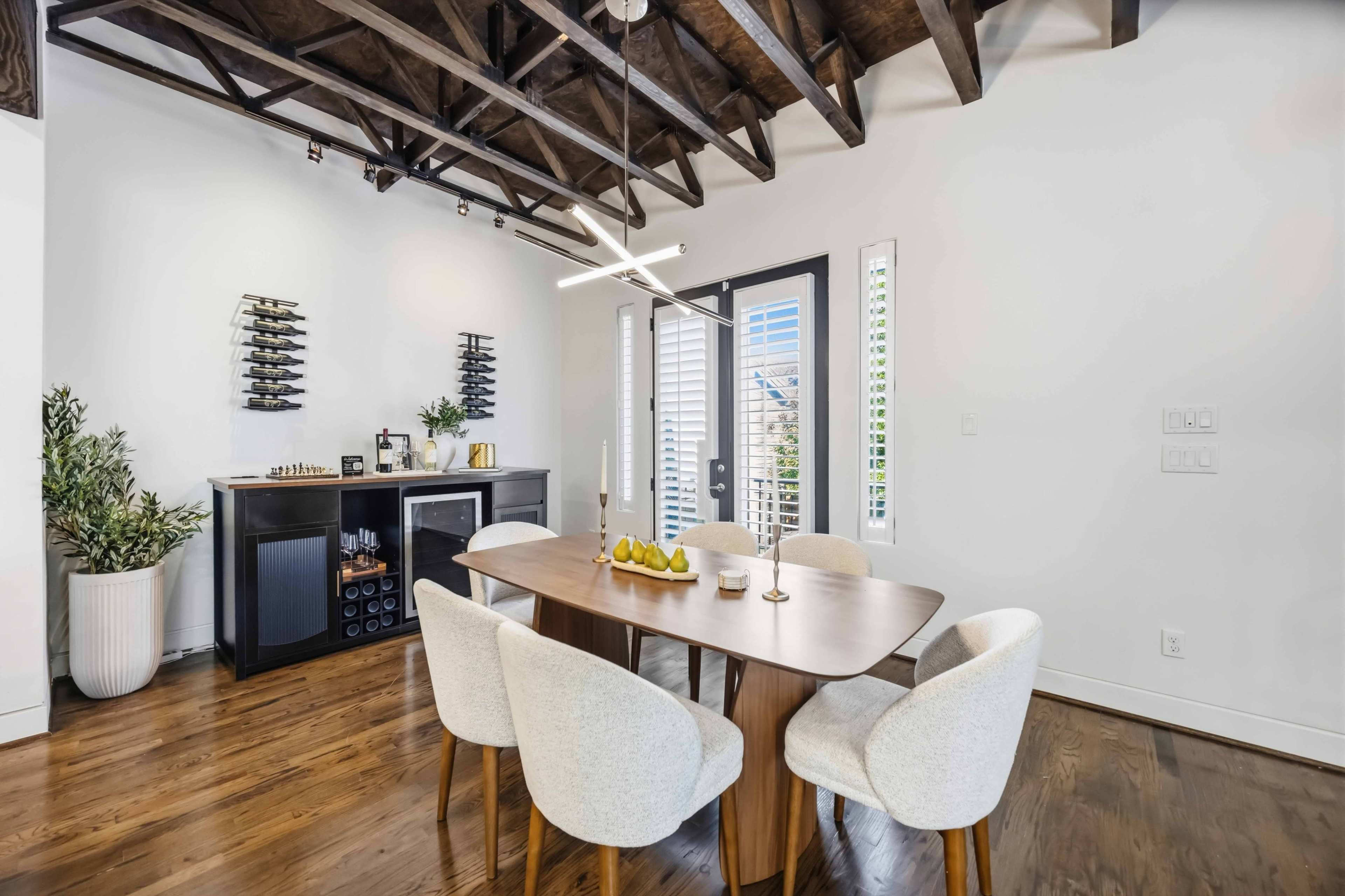 A modern dining area features a wooden table surrounded by upholstered chairs, with large windows and a wine rack on the wall.