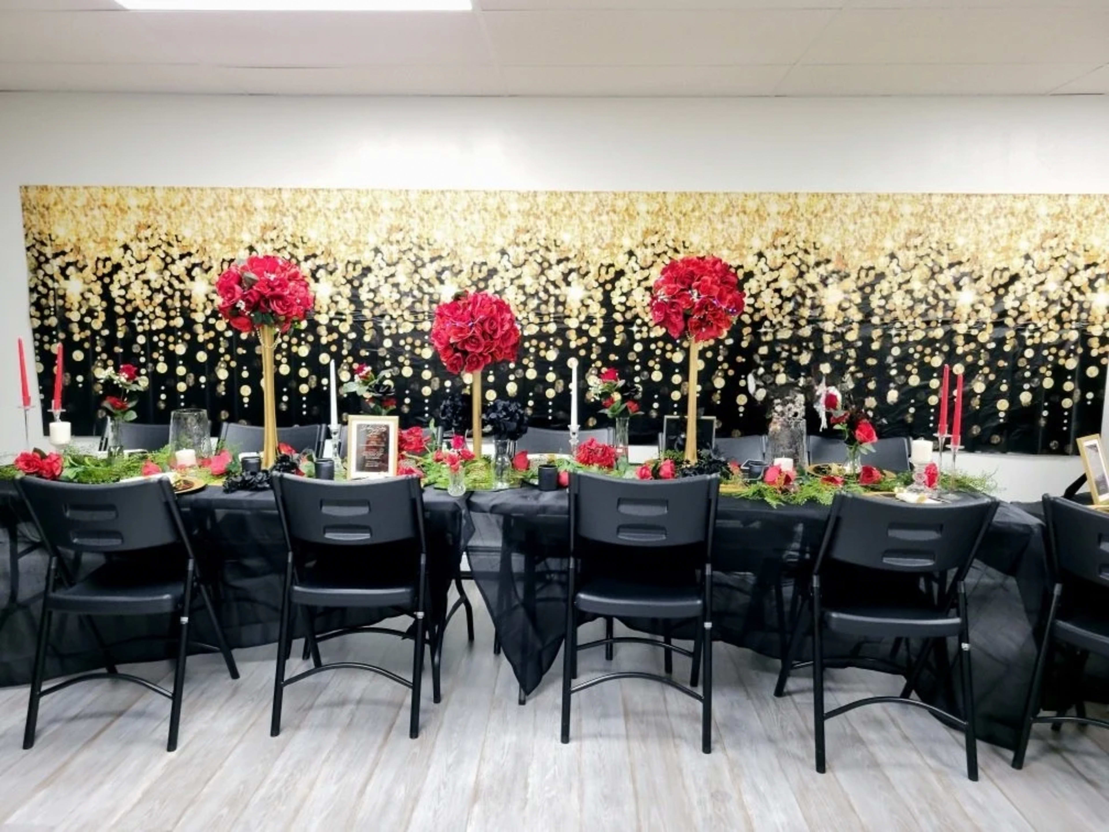 The image shows a beautifully arranged dining table with black tablecloths adorned with red floral centerpieces, candles, and decorative elements against a backdrop of a shimmering gold and black wall.