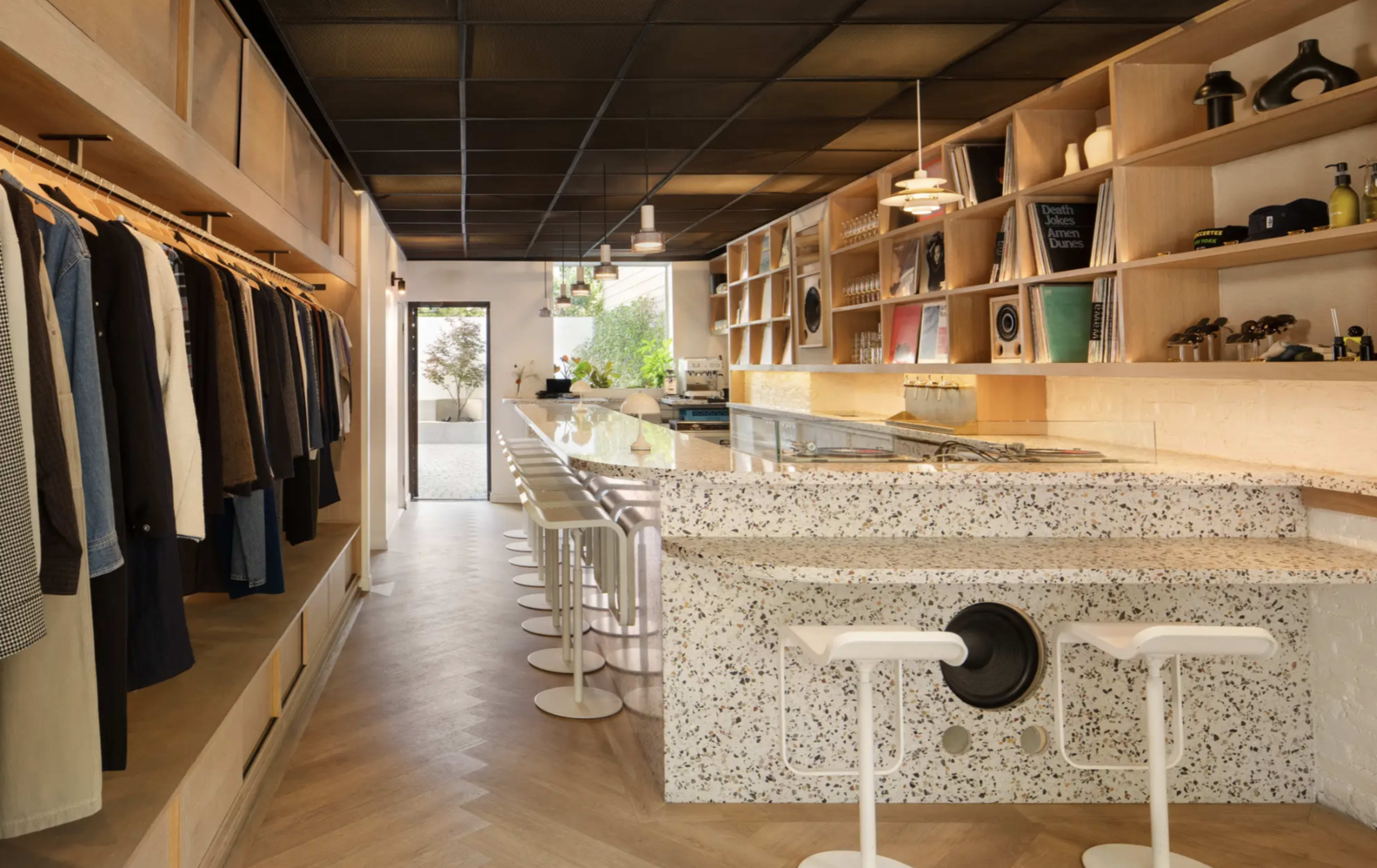 The image shows a modern retail space featuring a clothing display on one side and a minimalist bar area with white stools on the other.