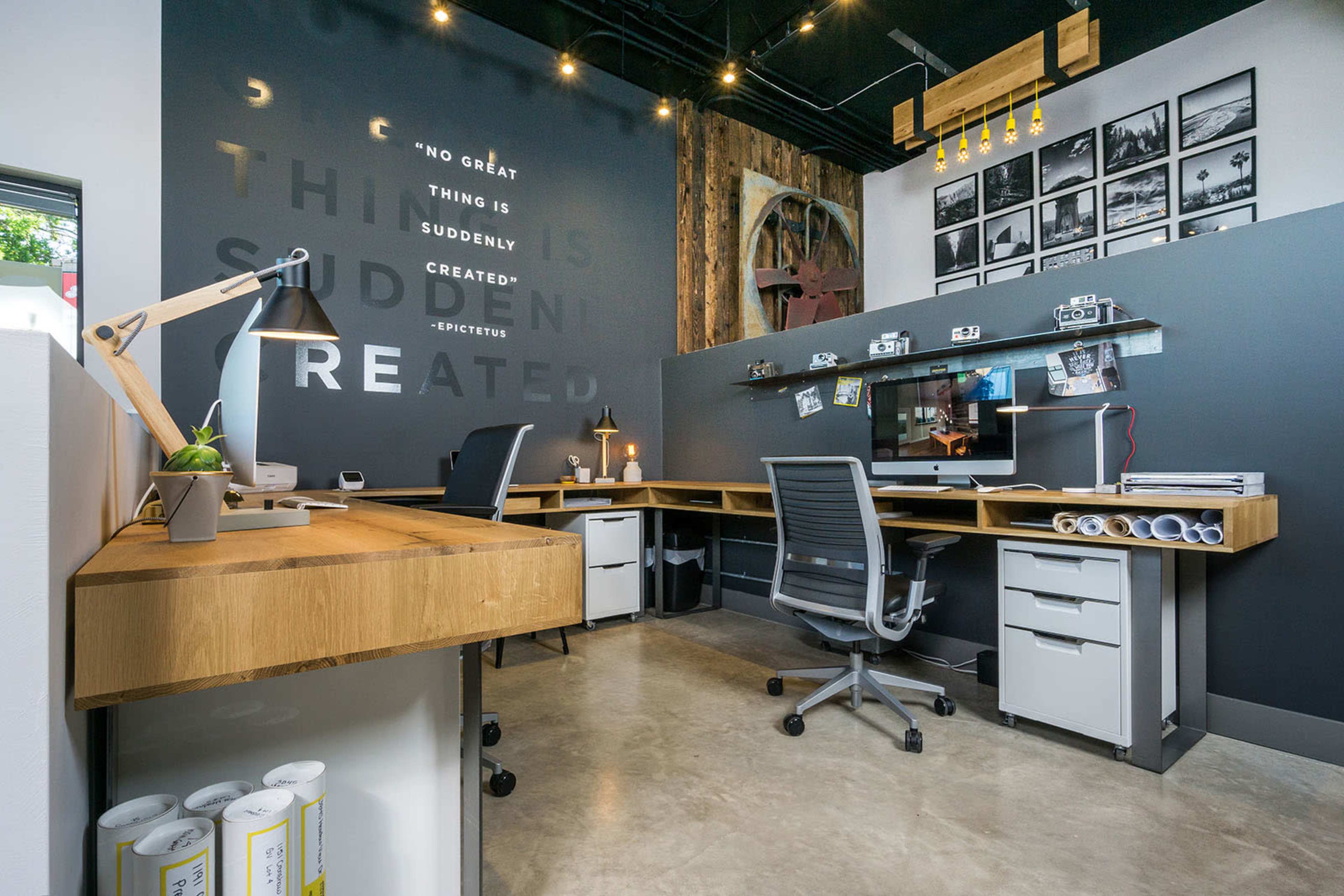 A modern office space with two desk workstations, wooden accents, and a dark wall featuring framed photographs and text.