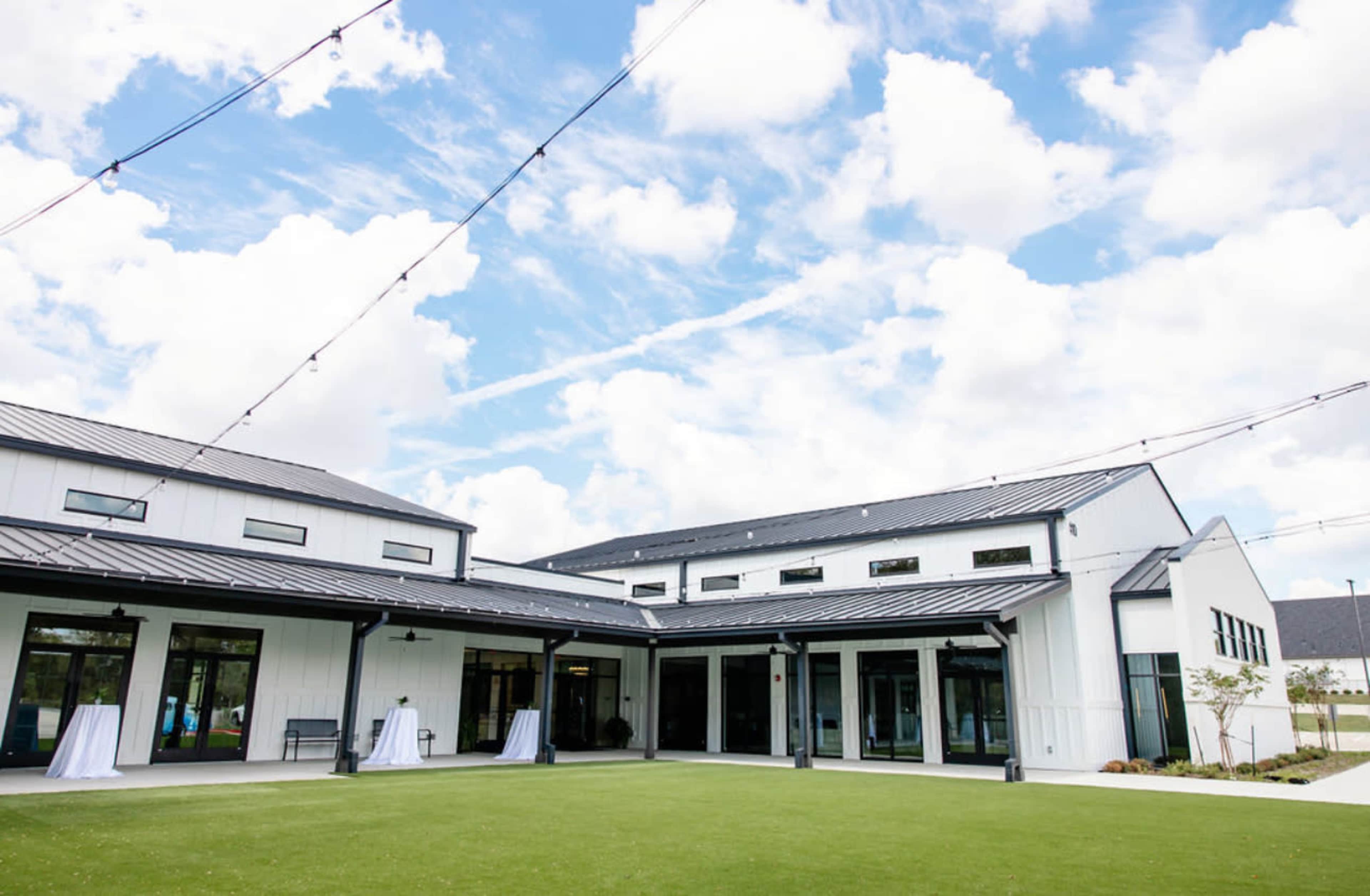 A modern building with a black metal roof and large glass doors is surrounded by a green lawn and outdoor string lights under a partly cloudy sky.