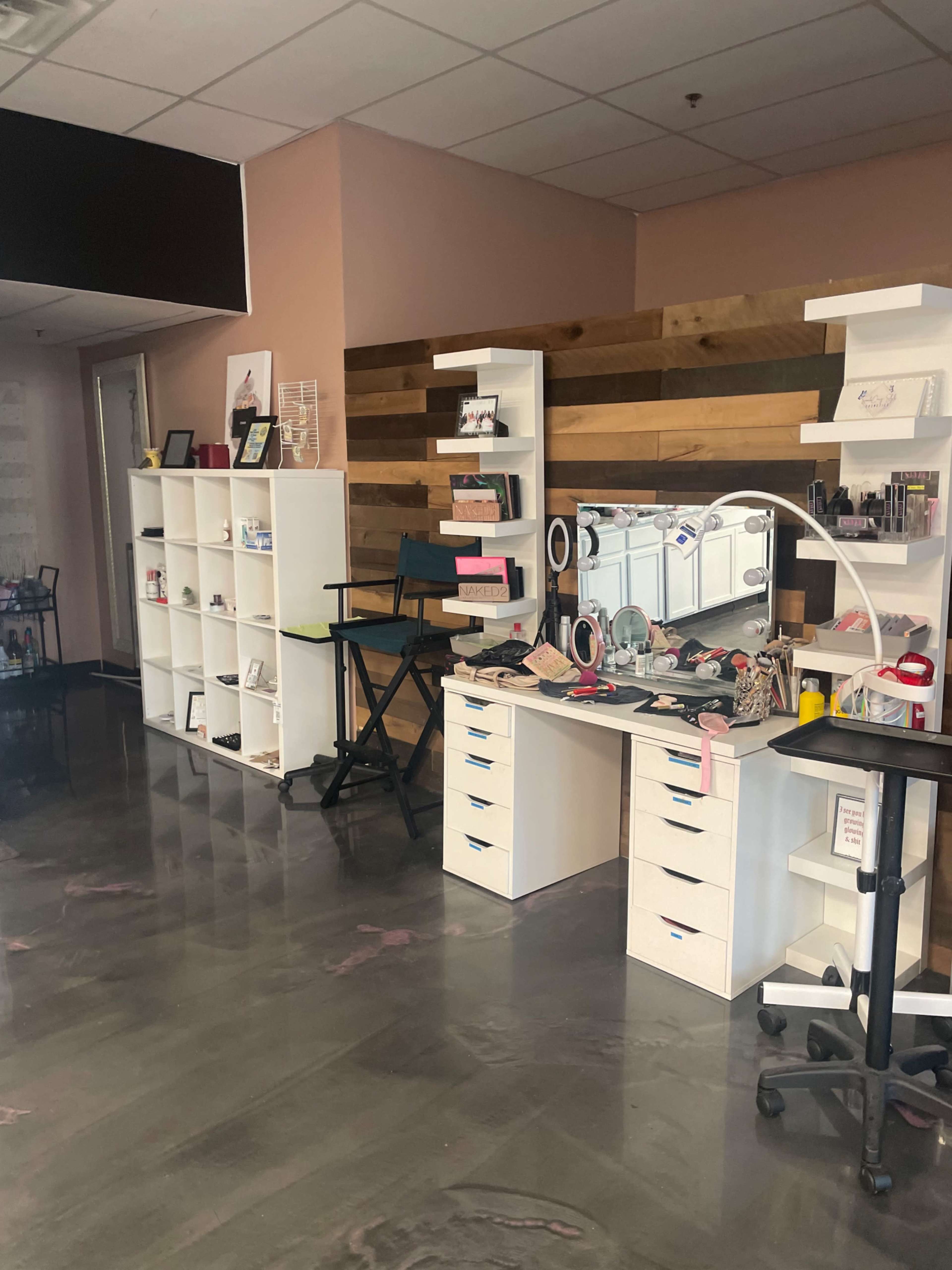 A beauty studio featuring a makeup station with a mirror, shelves containing cosmetics, and a chair beside a white storage unit.