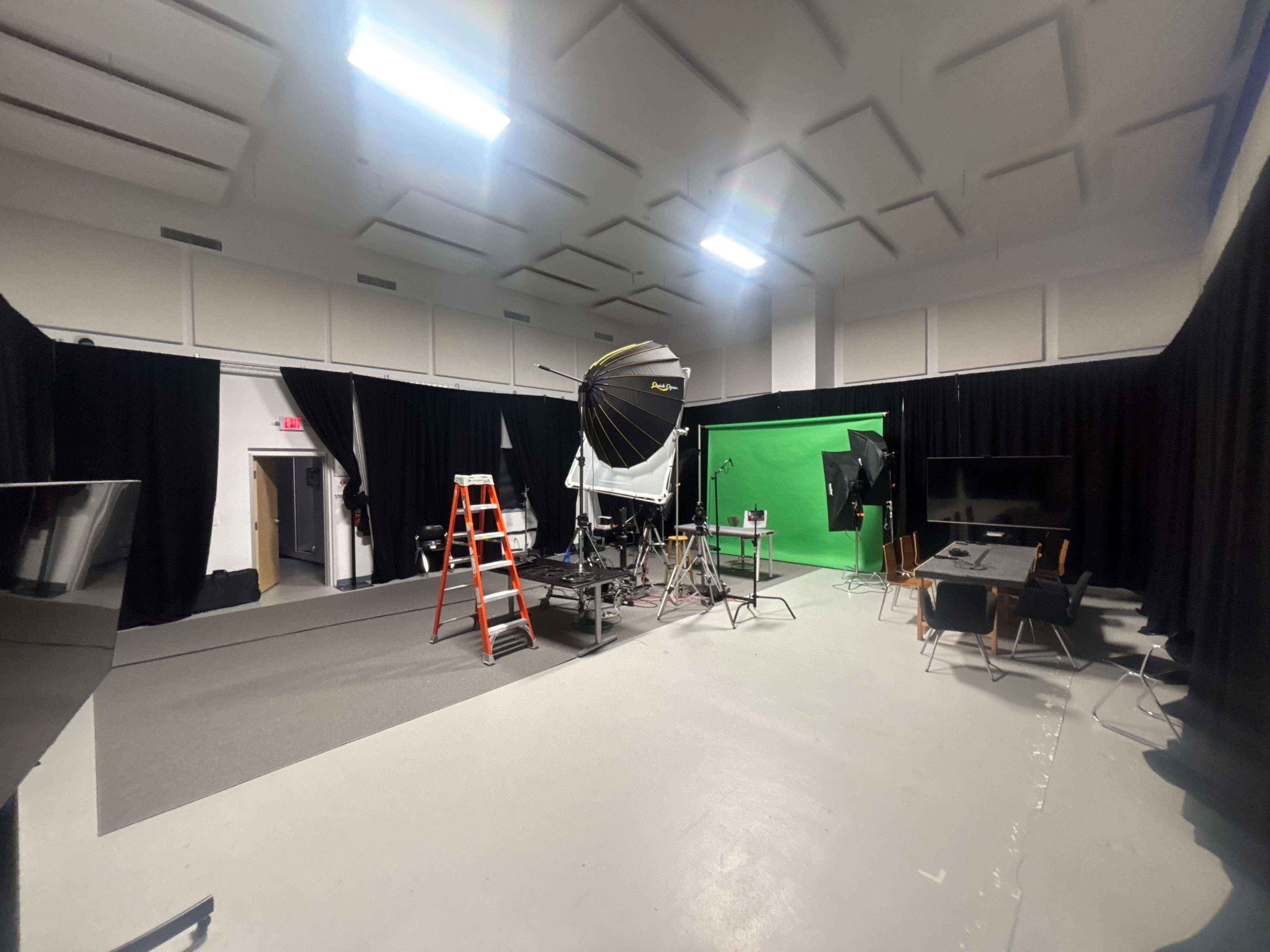 The image displays a photography studio equipped with a green screen, lighting setup, a ladder, and various pieces of equipment arranged on the floor.