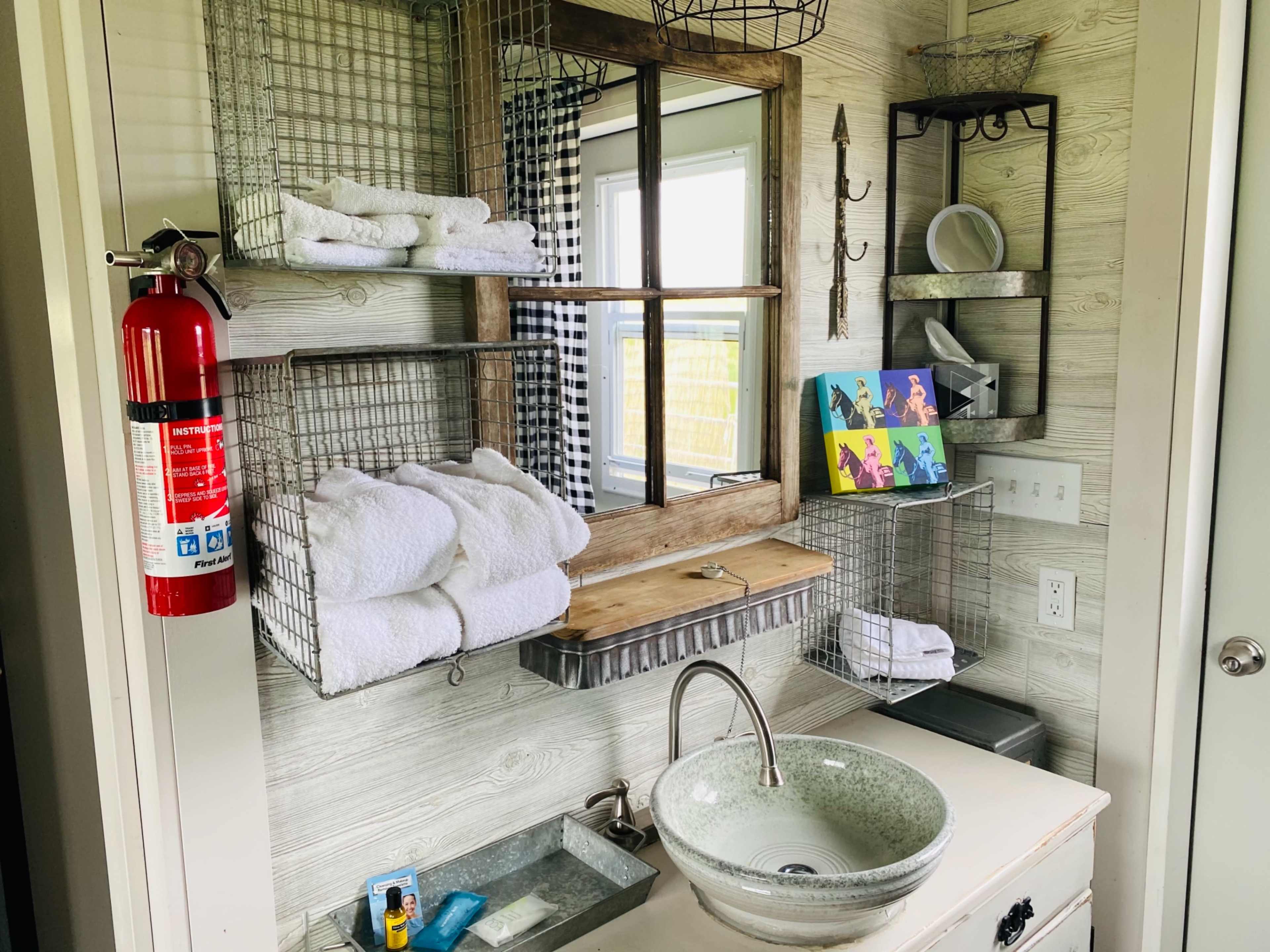 A bathroom featuring a sink, a wooden shelf with rolled towels, metal wire baskets for storage, a mirror, and a fire extinguisher mounted on the wall.