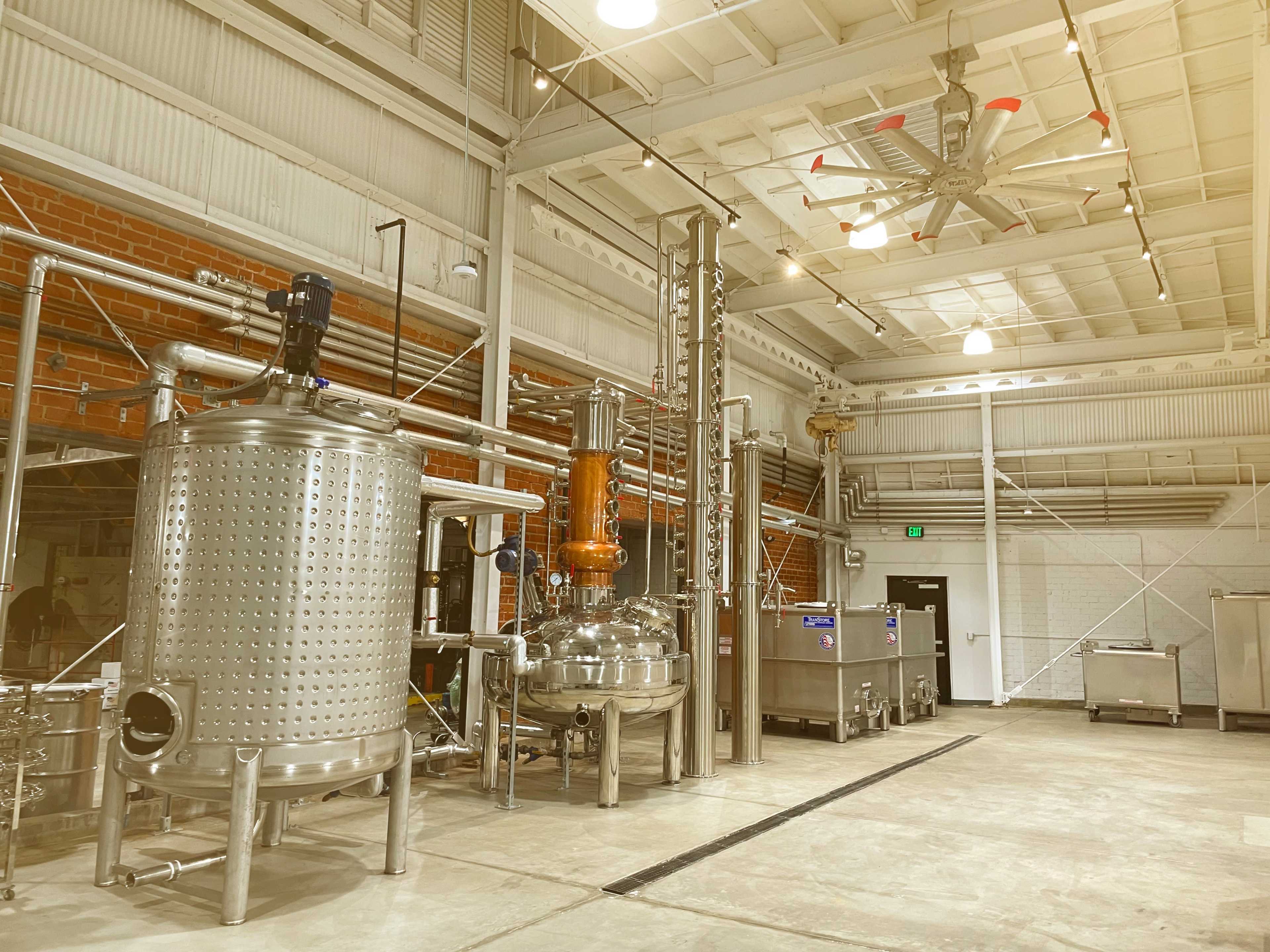 A spacious industrial distillery with stainless steel equipment, including fermentation tanks and a distillation column, against a backdrop of brick walls and overhead lighting.