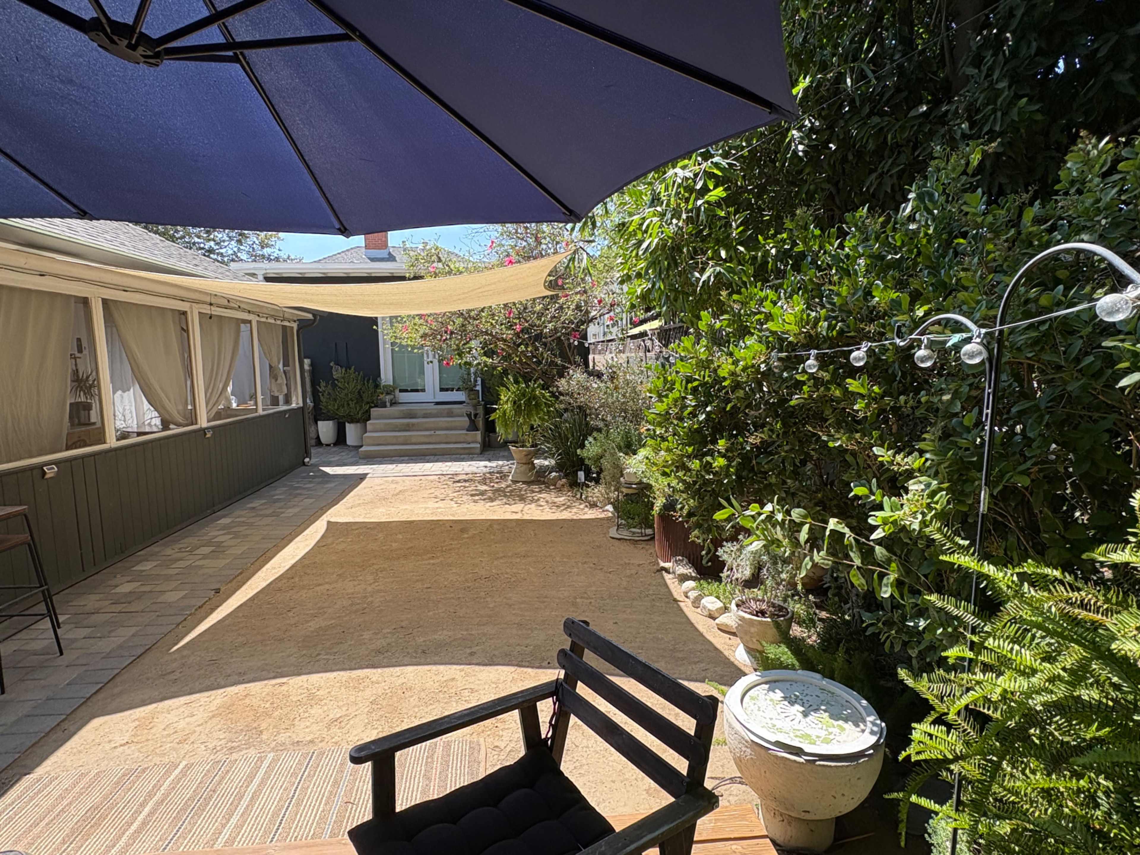 A shaded outdoor space features a chair, a sandy area, potted plants, and a pathway leading to a building.