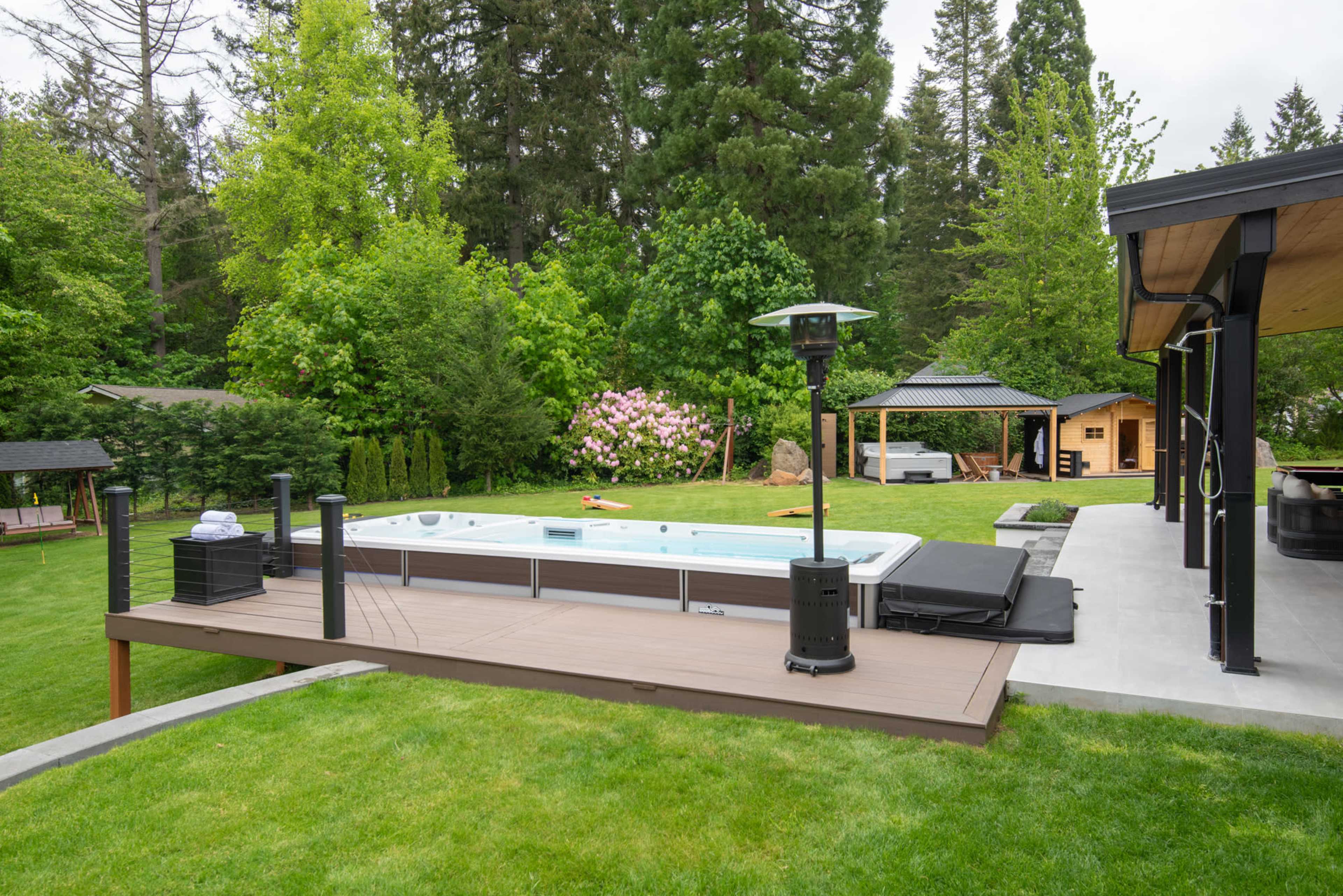 The image shows a landscaped backyard with a hot tub, surrounded by greenery and outdoor features like a gazebo and patio heaters.