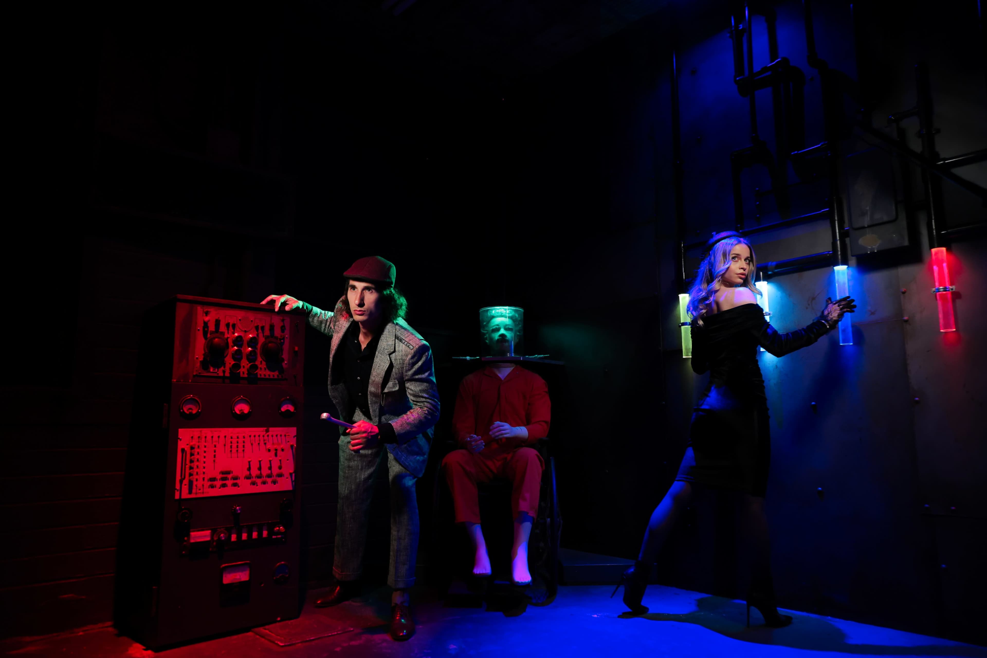 A man in a suit and hat stands beside a vintage control panel, while a woman in a black dress poses with glowing tubes, and a figure in a red outfit sits silently in the background.