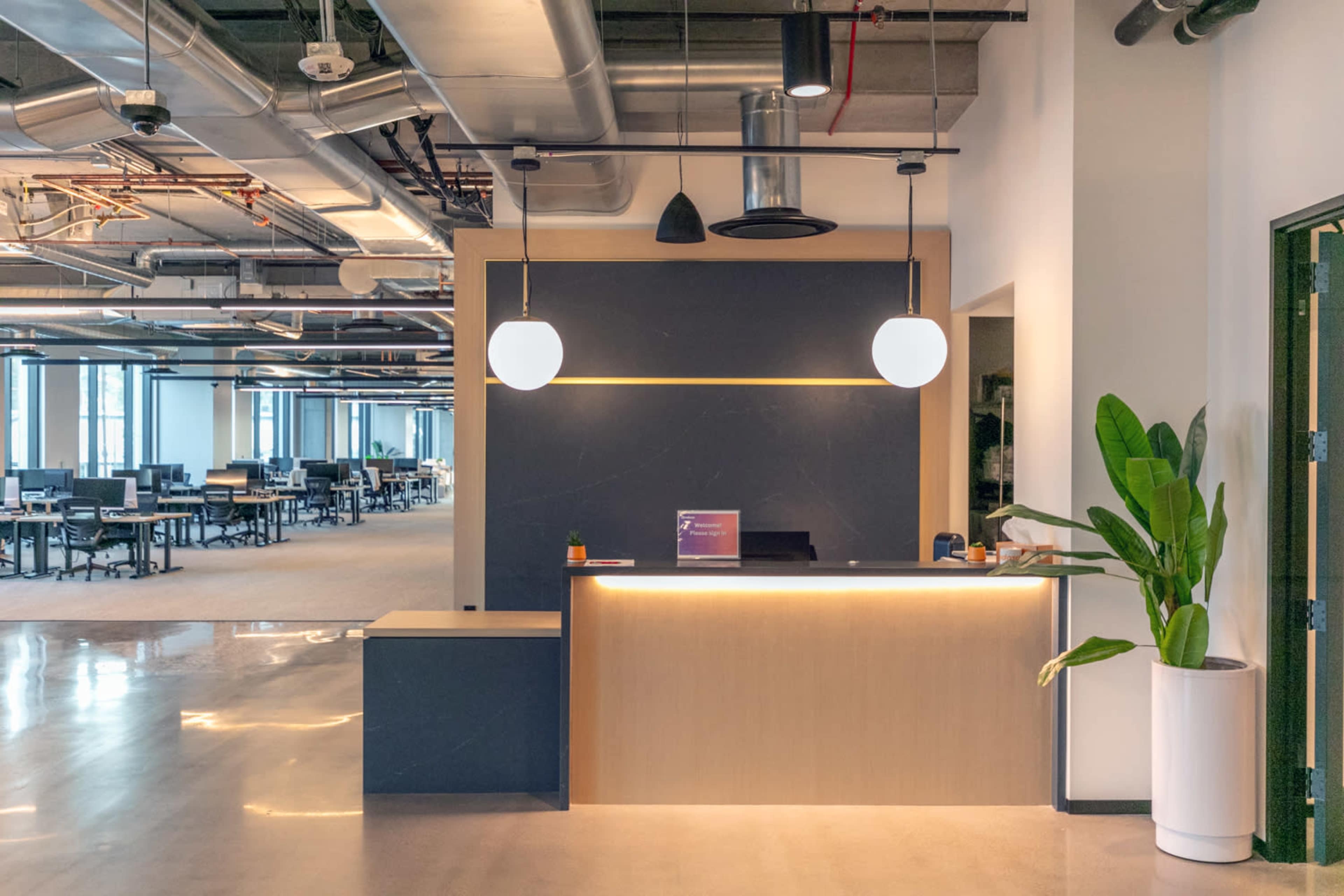 A modern office reception area with a minimalist front desk, overhead lighting, and a potted plant beside it, set against an open workspace with rows of desks in the background.