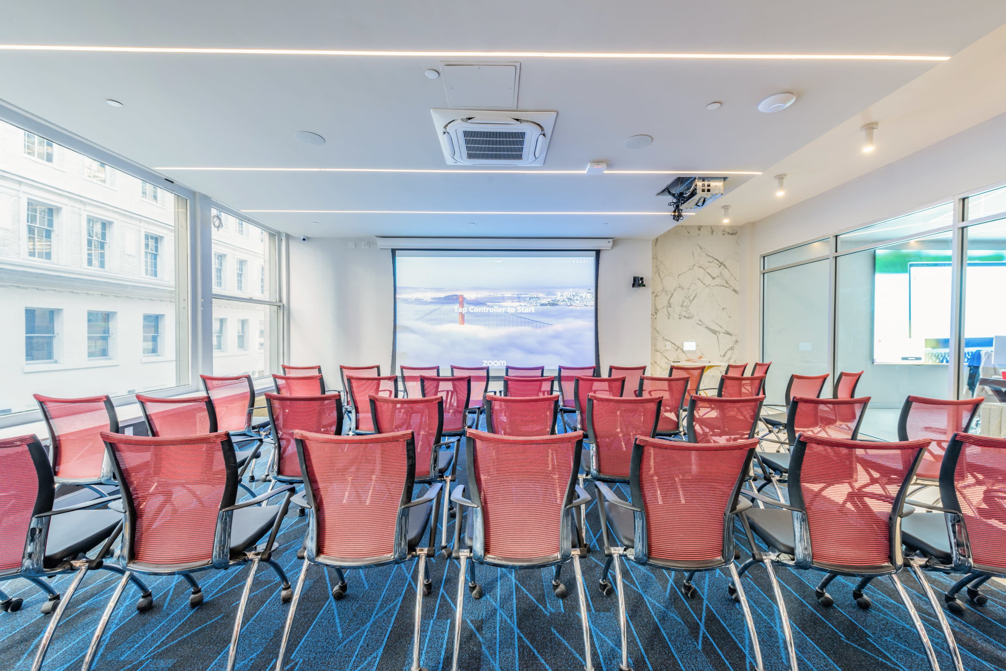The image shows a modern conference room with rows of red mesh chairs facing a projection screen.