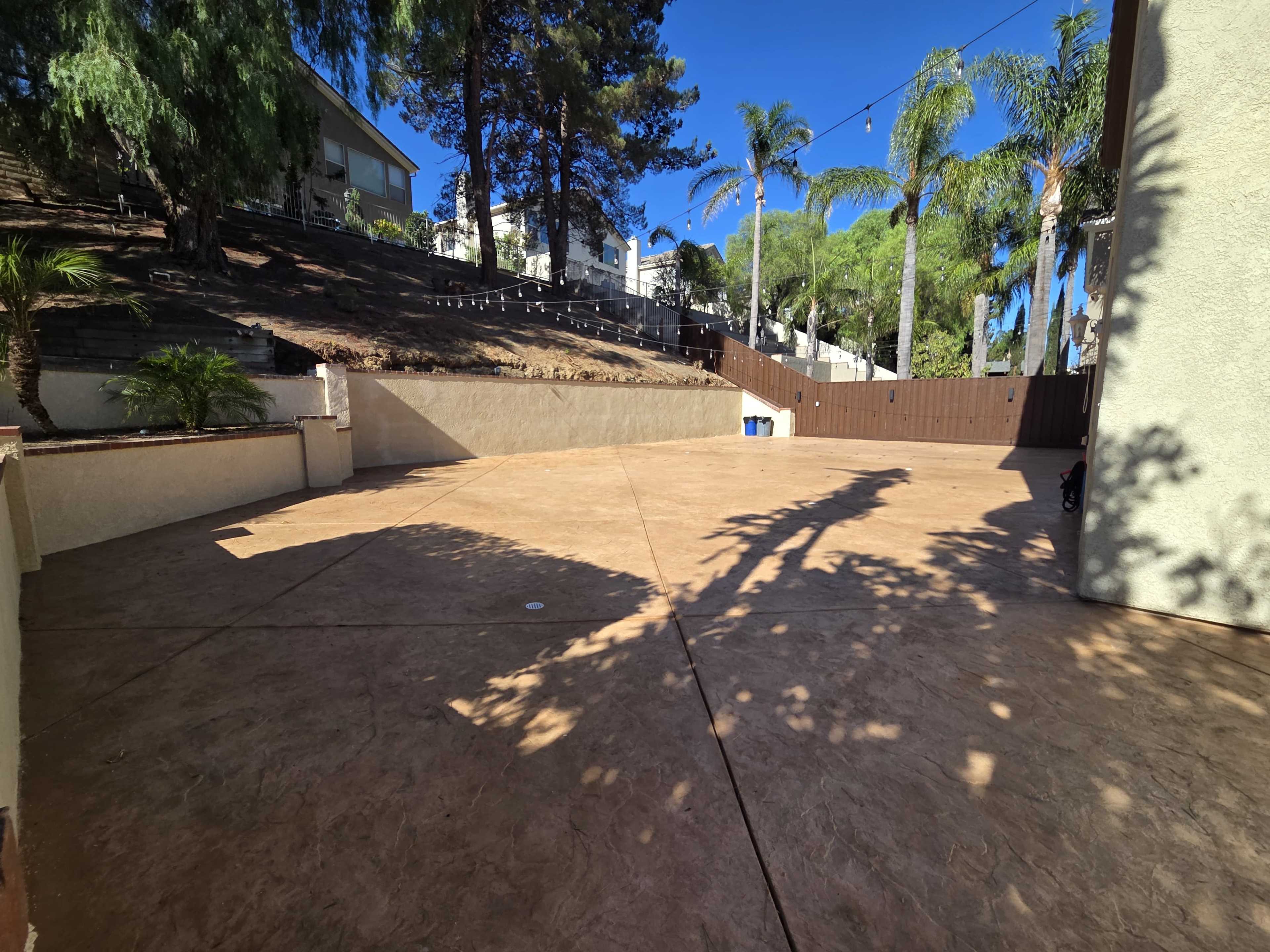 The image shows a spacious concrete patio area surrounded by trees and houses, with a brown fence in the background.