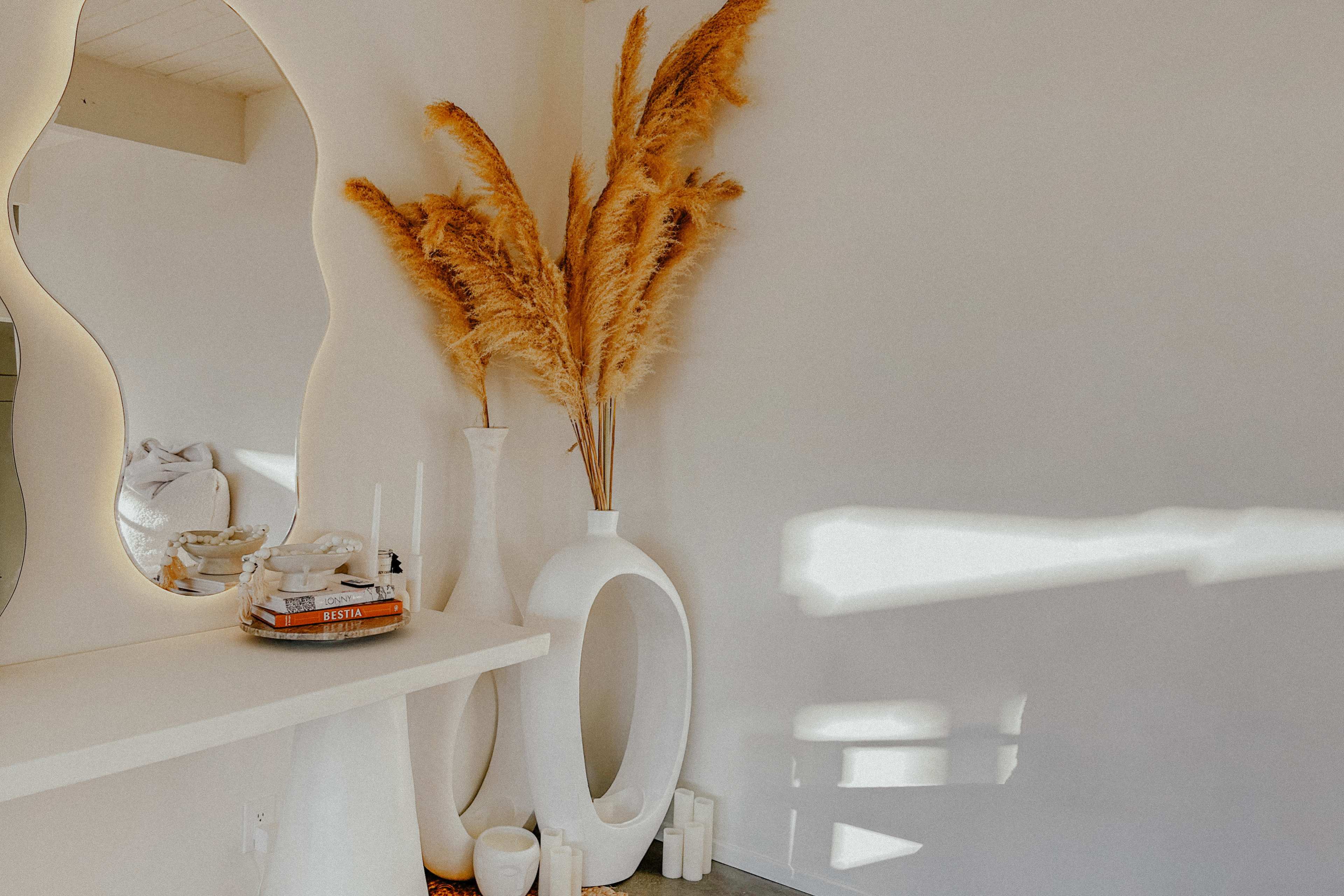 A minimalistic interior space featuring a large mirror, decorative pampas grass in a vase, and various candles arranged on a table against a white wall.