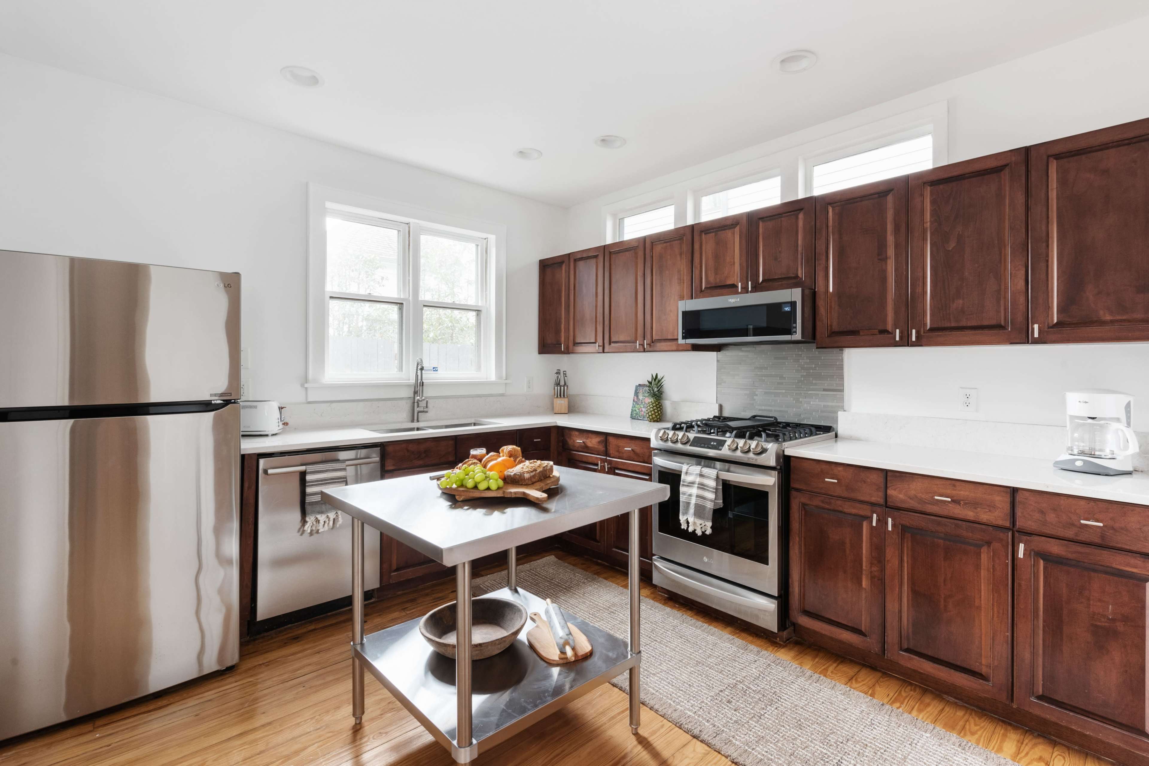 A kitchen with wooden cabinets, a stainless steel refrigerator, and a central island with fruits and a bowl, illuminated by natural light from the windows.
