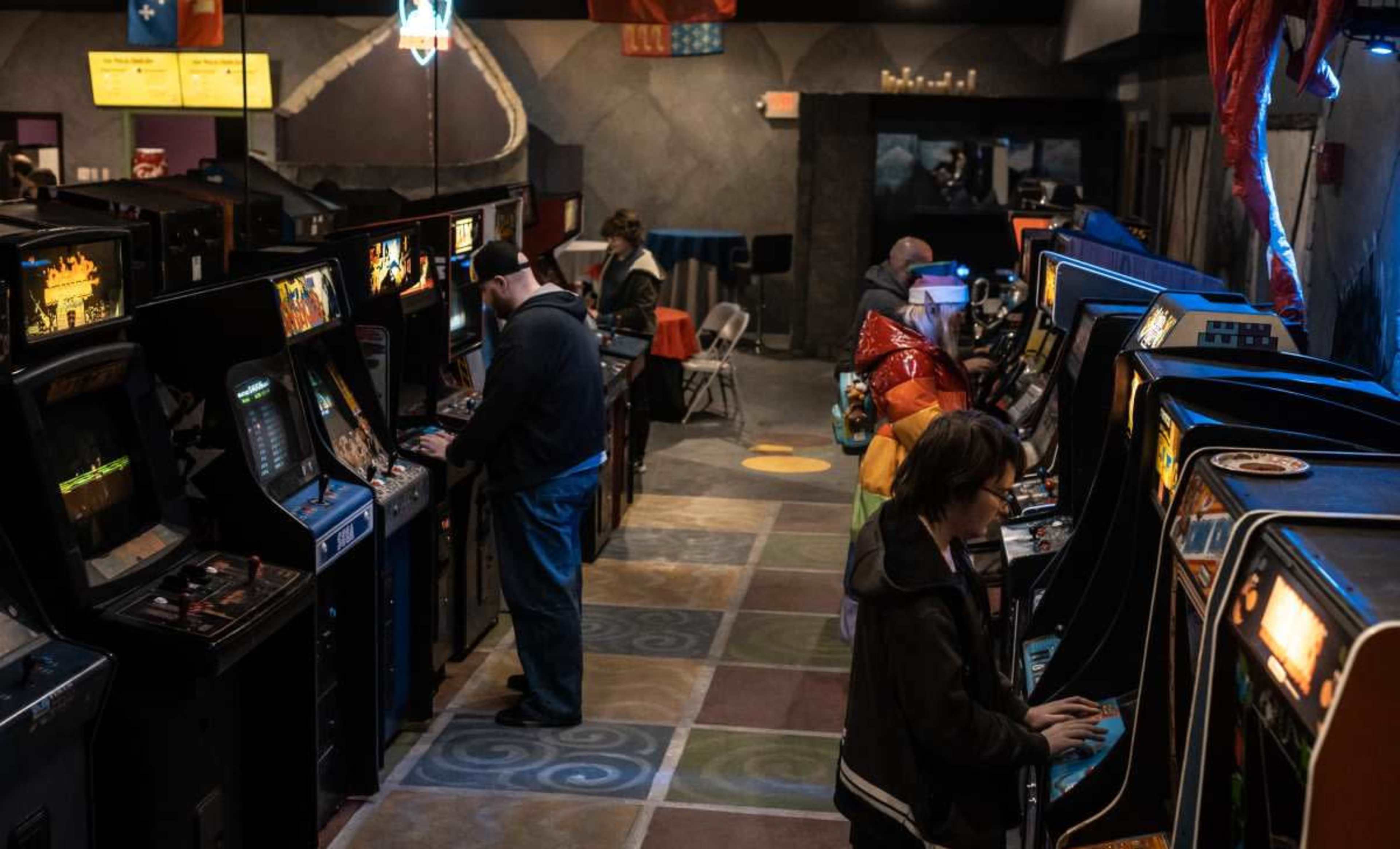 The image shows a retro arcade with several arcade game machines, where people are playing games and others are walking around in a dimly lit environment.