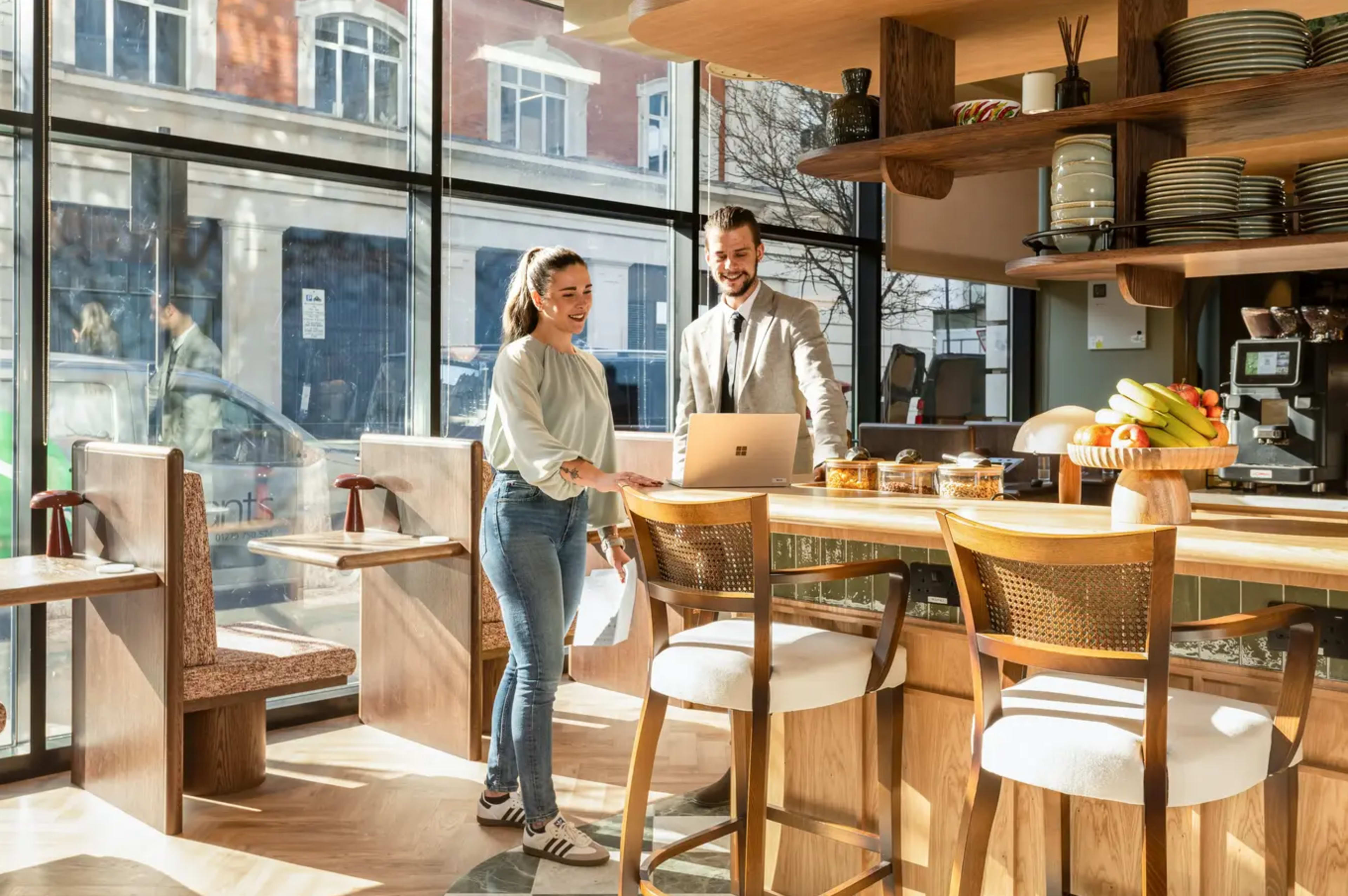 A woman and a man are engaged in conversation while working on a laptop at a wooden countertop in a cafe with large windows.