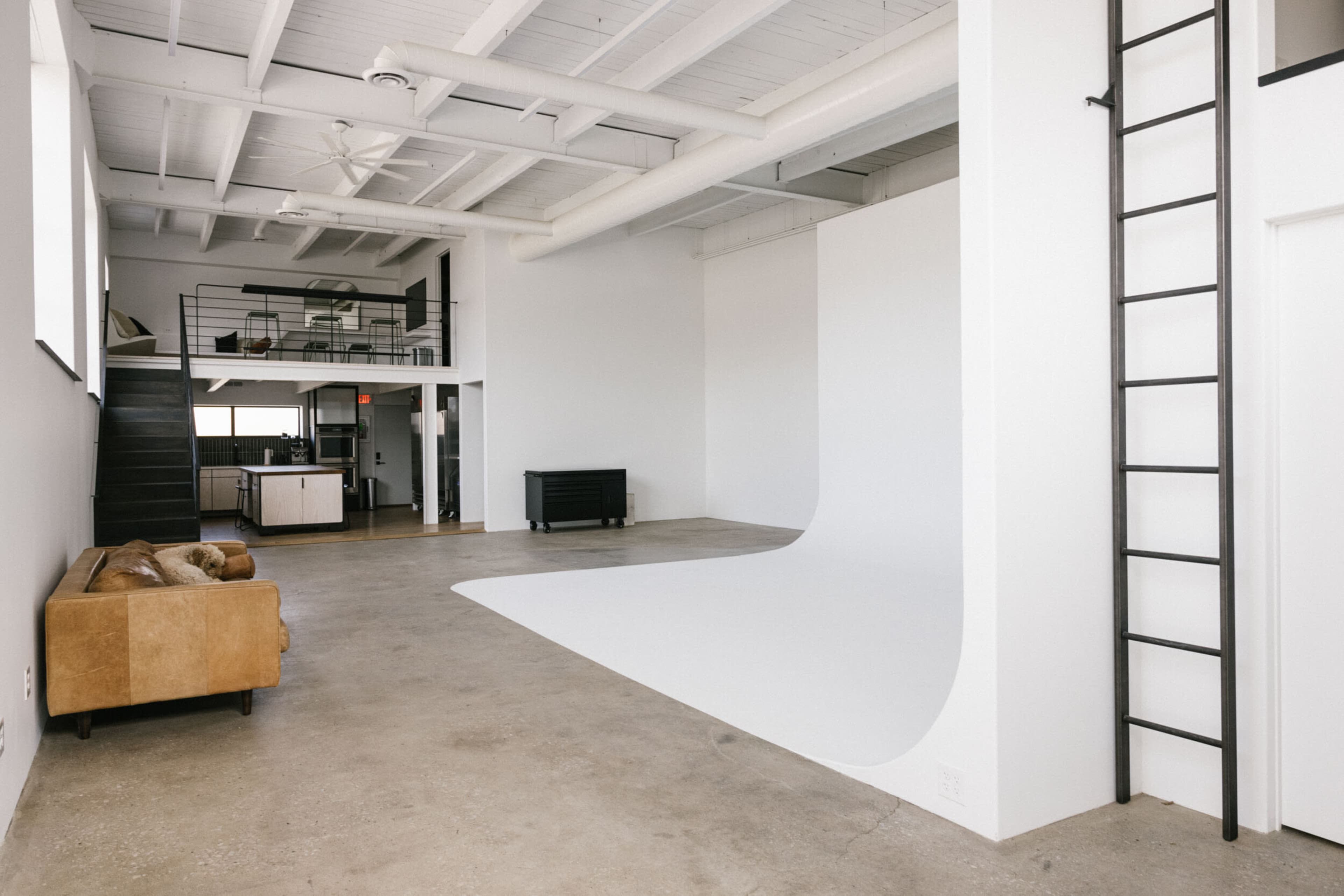 A spacious, minimalistic studio with a curved white backdrop, a brown sofa, and a staircase leading to an upper level.