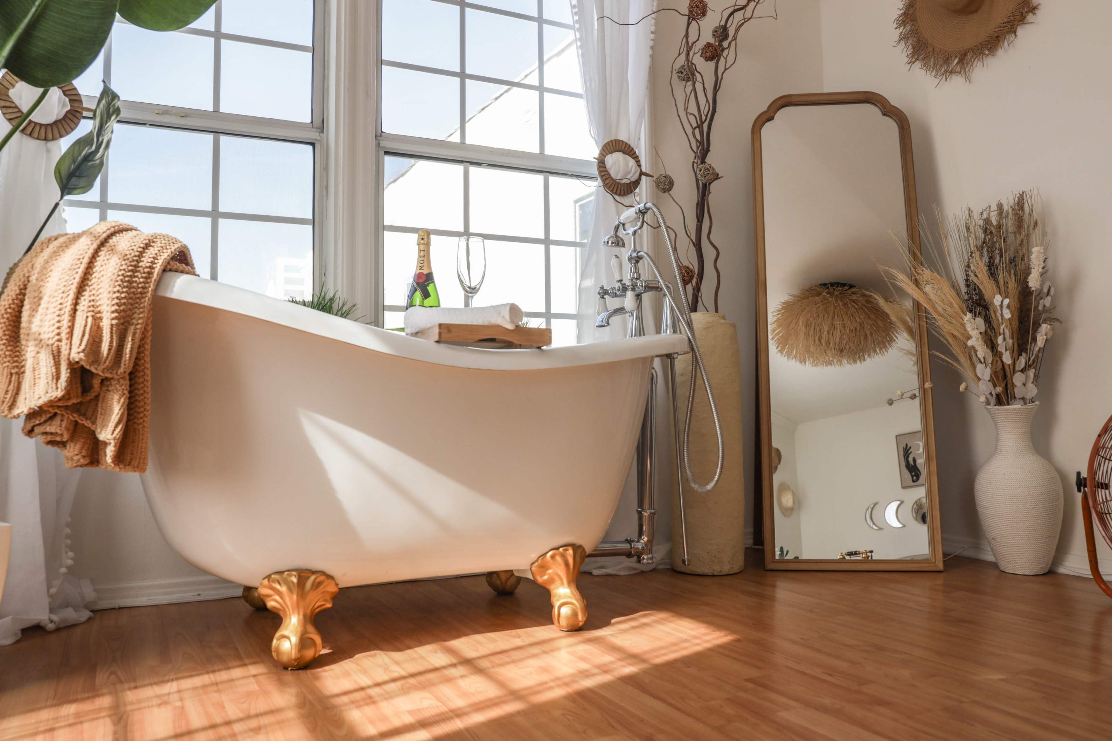 A clawfoot bathtub is positioned beside a large window, with a tall mirror and decorative plants nearby.