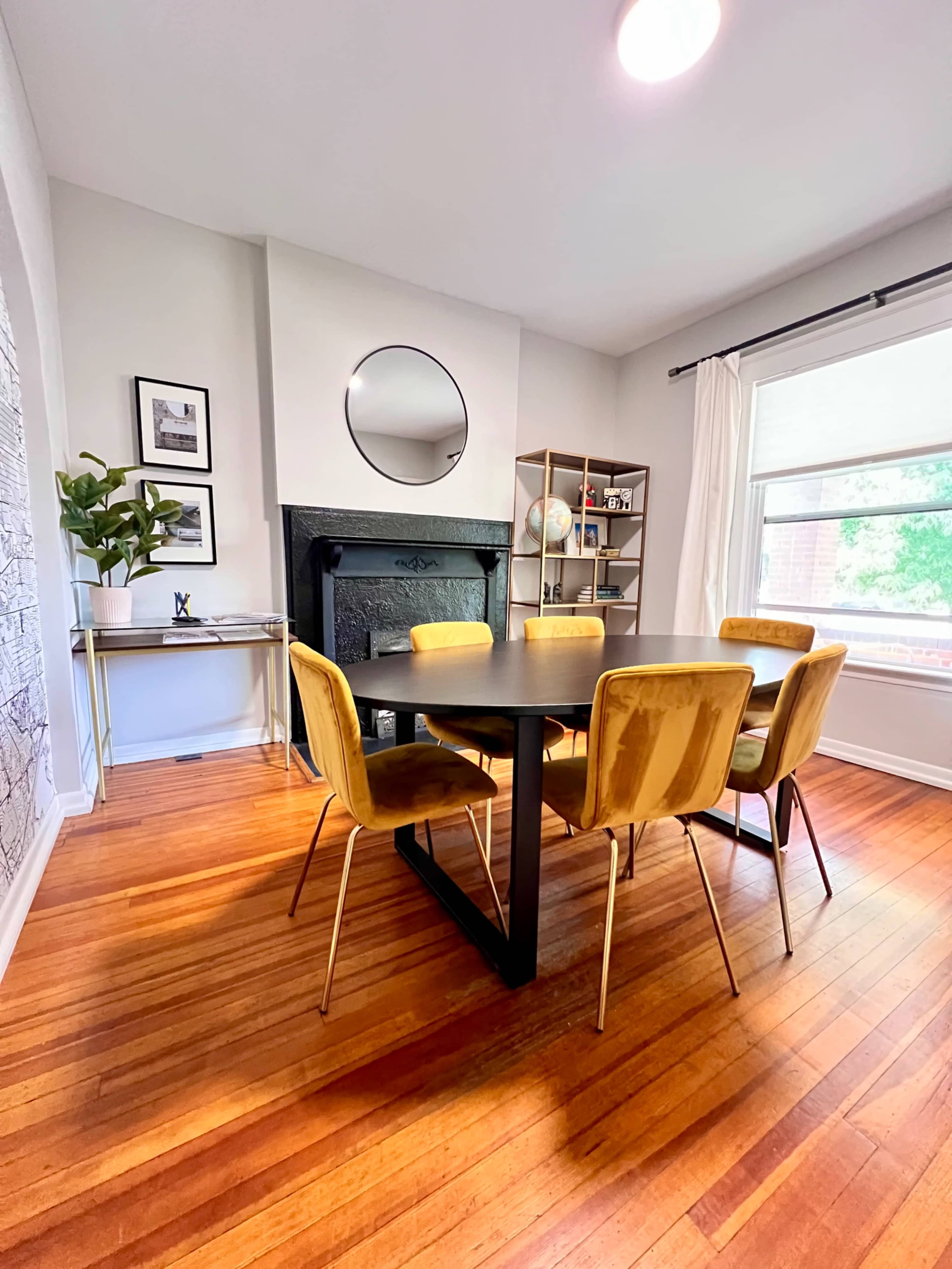 The image shows a dining area featuring a wooden table surrounded by six yellow chairs, with a mirror and a bookshelf on the wall.