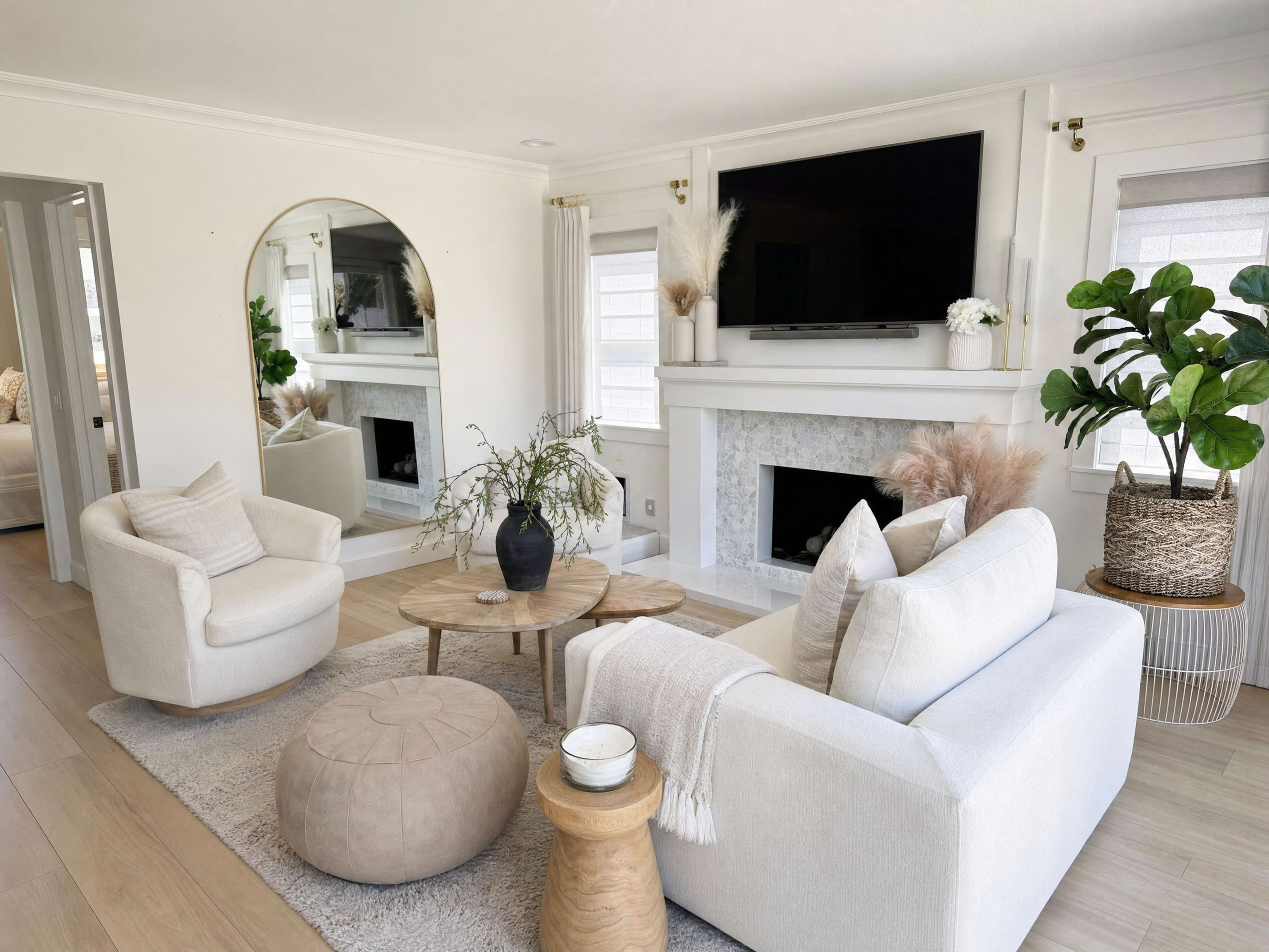 The image shows a bright and modern living room with a fireplace, two light-colored armchairs, a round coffee table, and indoor plants, all arranged on light wood flooring.