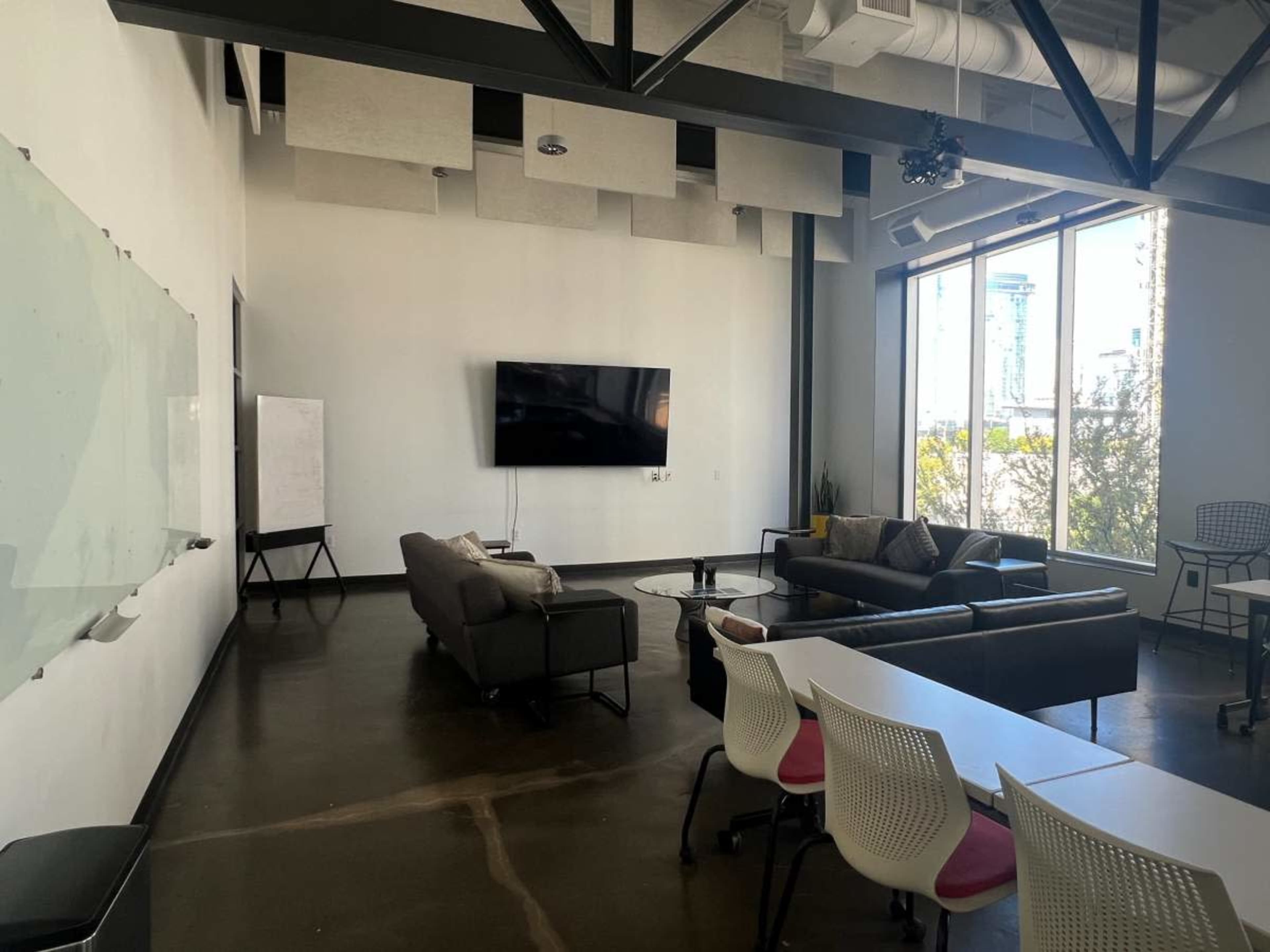 A modern office space features a large window, a television mounted on the wall, a seating area with a couch, and a long table with chairs.