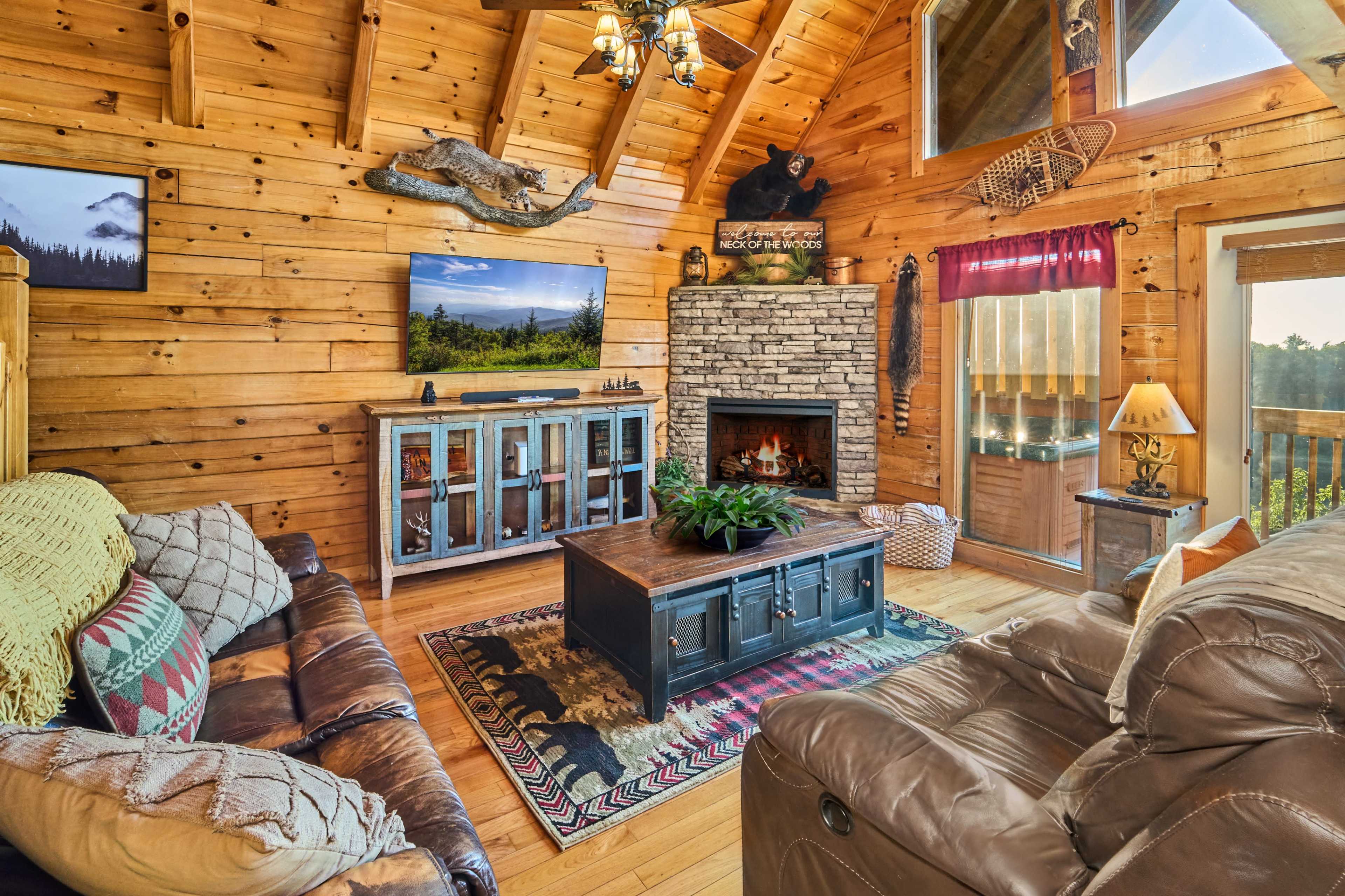 The image shows a cozy wooden cabin living room with a stone fireplace, a large television, and comfortable seating arranged around a coffee table.