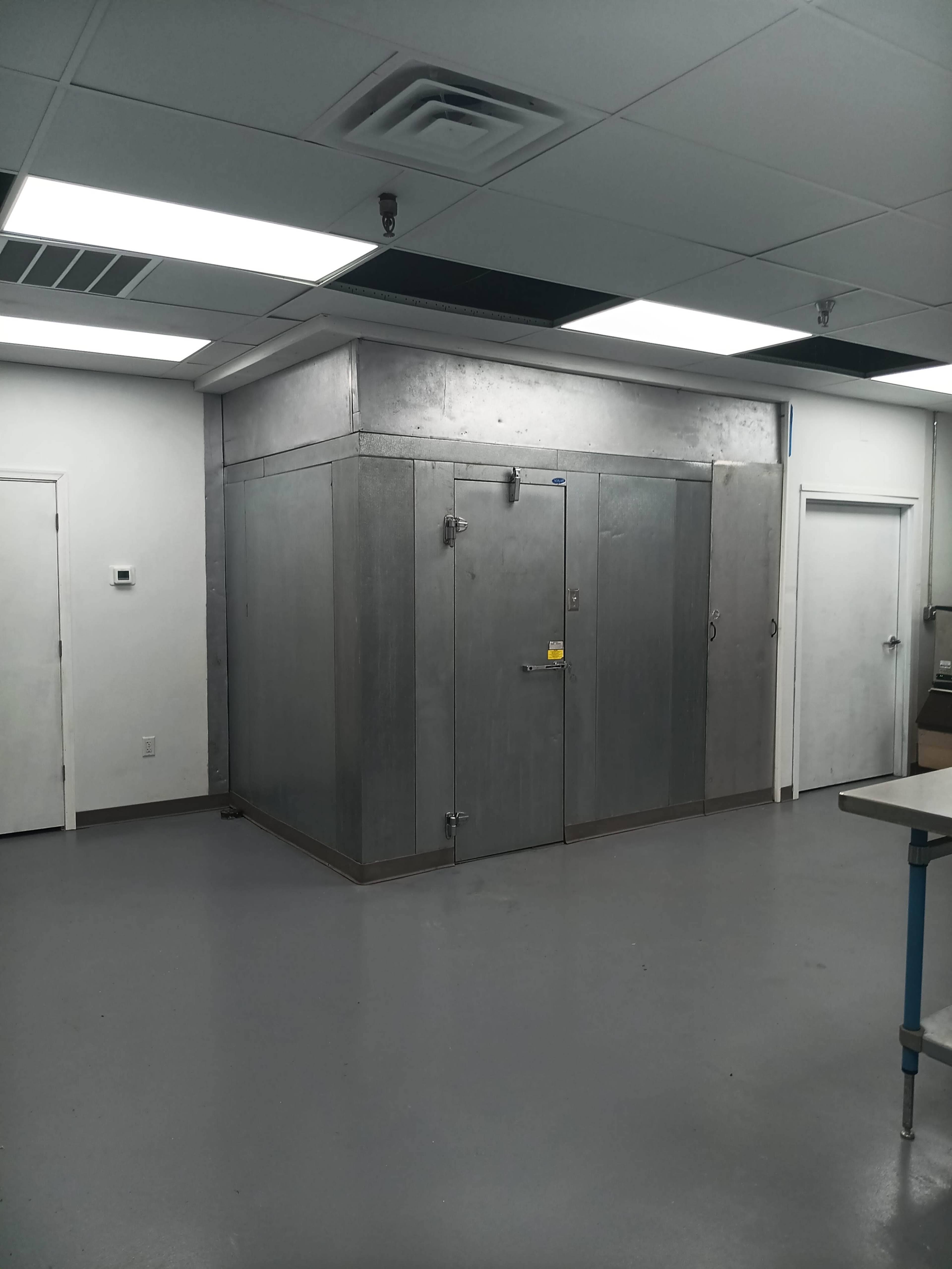 The image shows a large metal walk-in cooler with a double-door entrance, situated in a well-lit room with white walls and a gray floor.