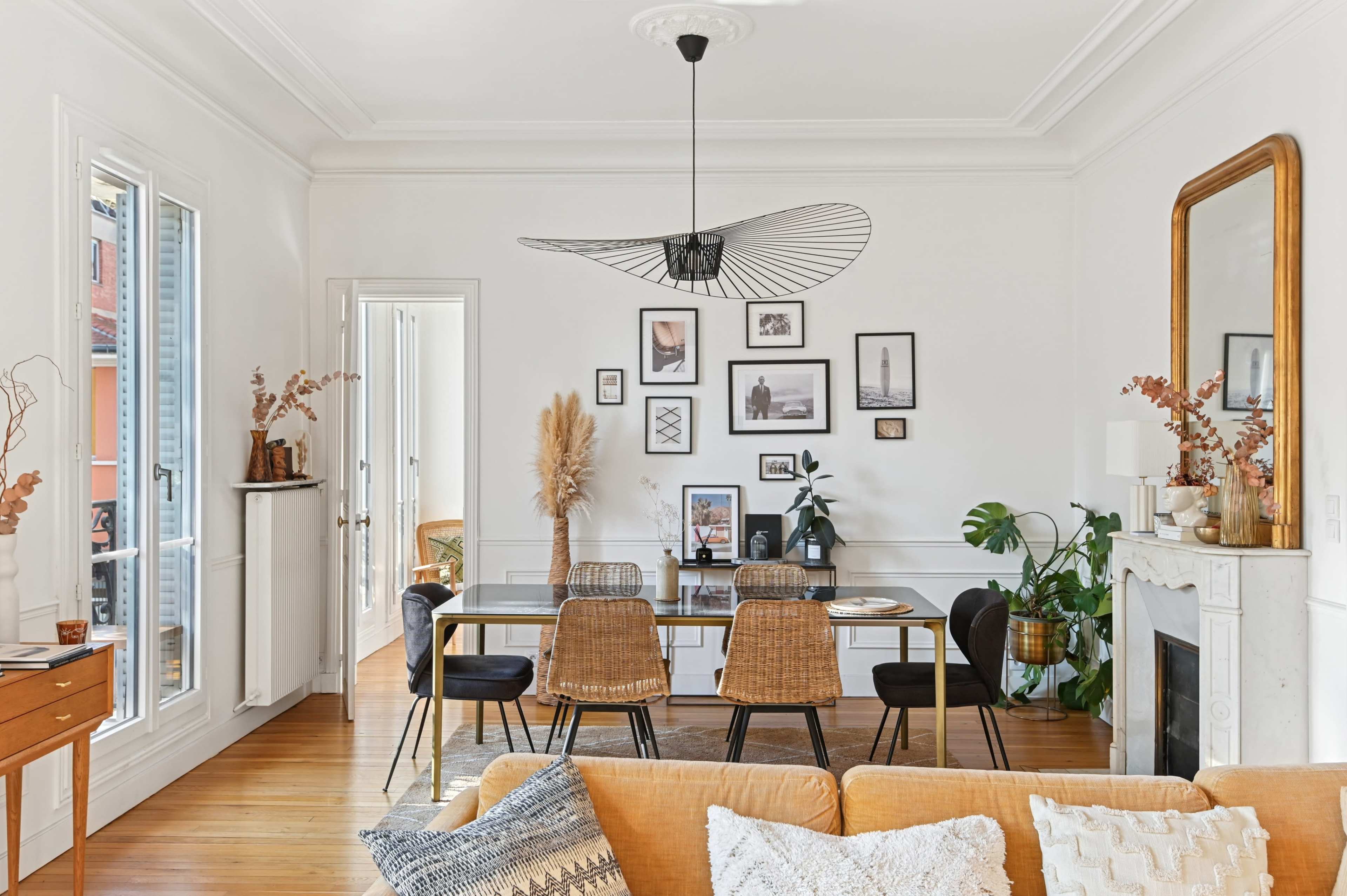 A modern dining area features a black table surrounded by woven chairs, framed photos on the wall, and a large mirror above a white fireplace.