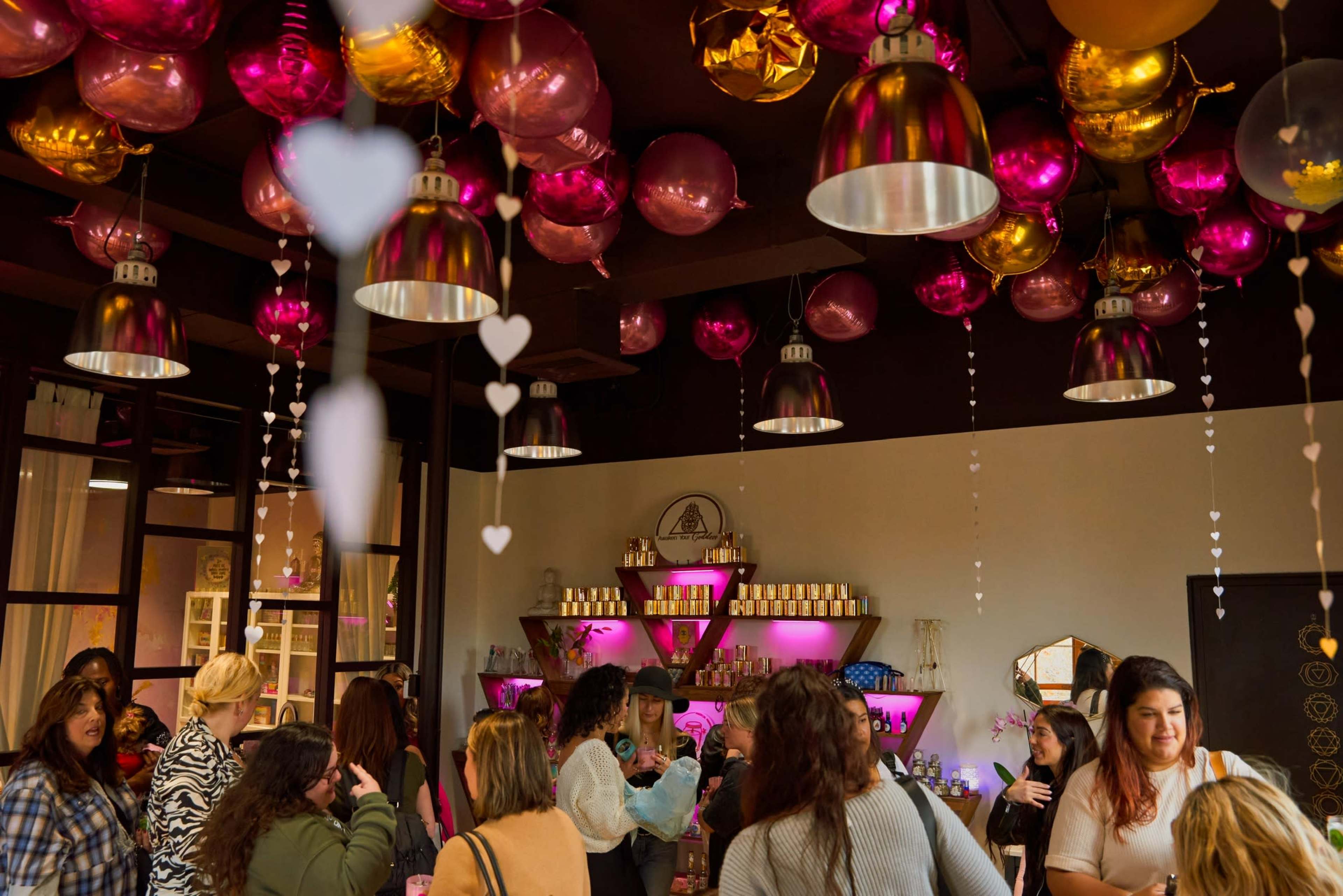 A vibrant gathering takes place in a room decorated with pink and gold balloons, featuring pendant lights and shelves filled with jars and decorative items.