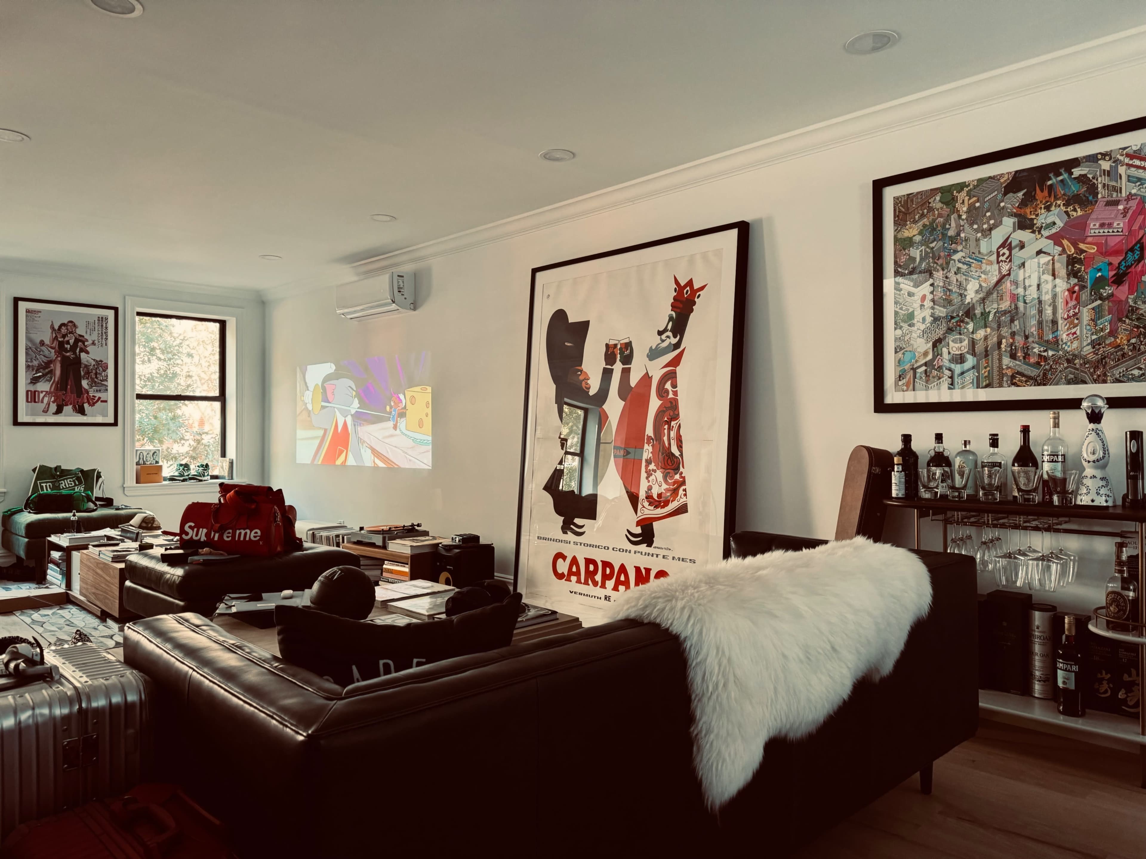 A modern living room featuring a black leather couch, various art pieces on the walls, and several items like luggage and clothing scattered around.