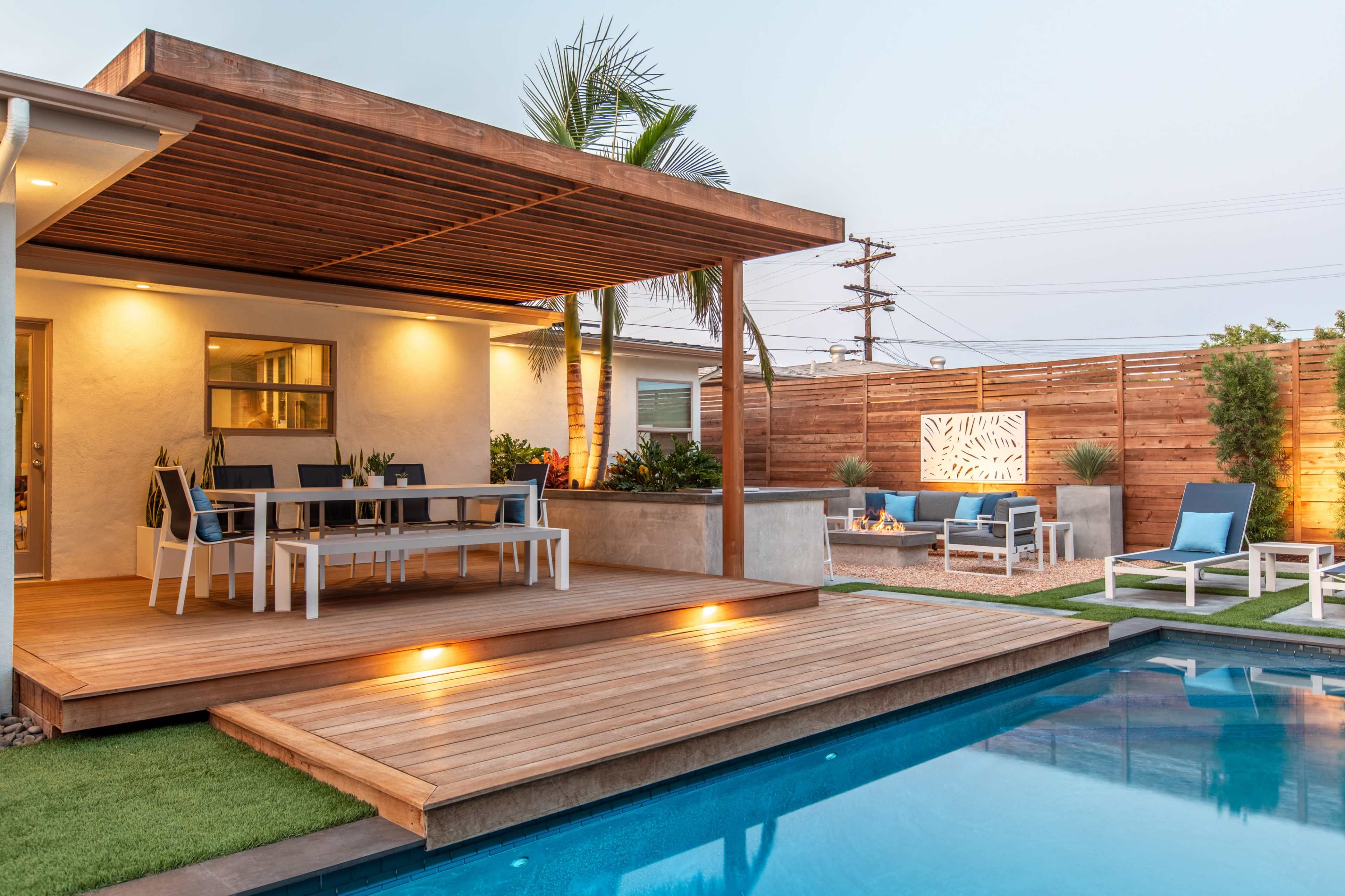 A modern outdoor patio features a dining table beneath a wooden pergola, adjacent to a rectangular pool surrounded by landscaped greenery.
