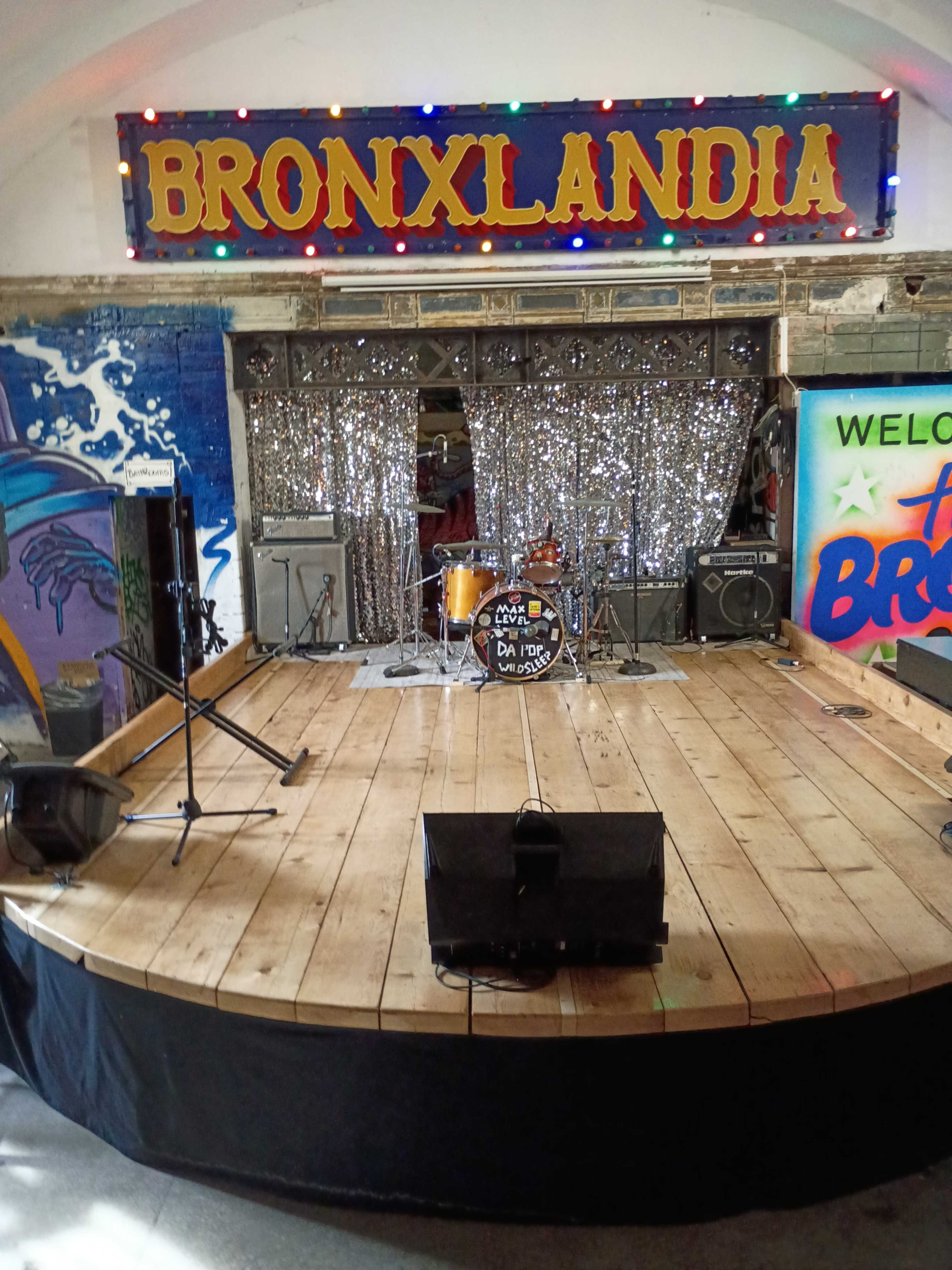 The image shows a stage at Bronxlandia, featuring a wooden floor, colorful lights, musical instruments, and a backdrop with glittering decorations.
