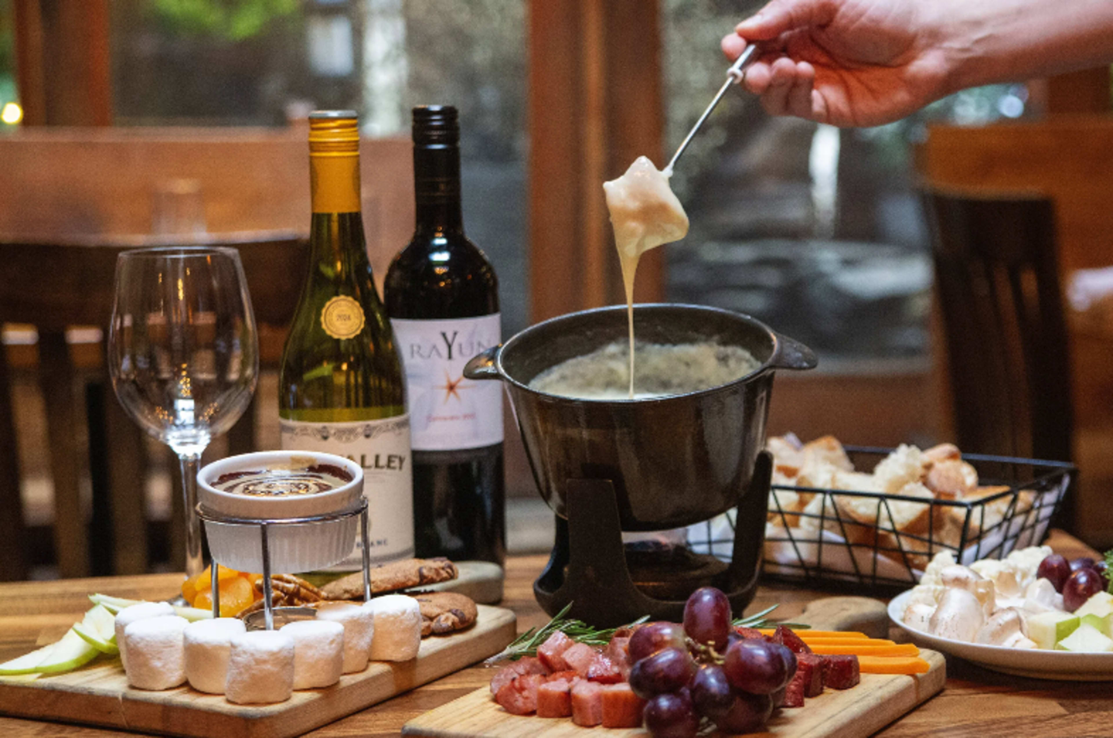 A hand holds a skewer with a piece of food over a pot of melted cheese, surrounded by a charcuterie board, wines, and various dipping items.