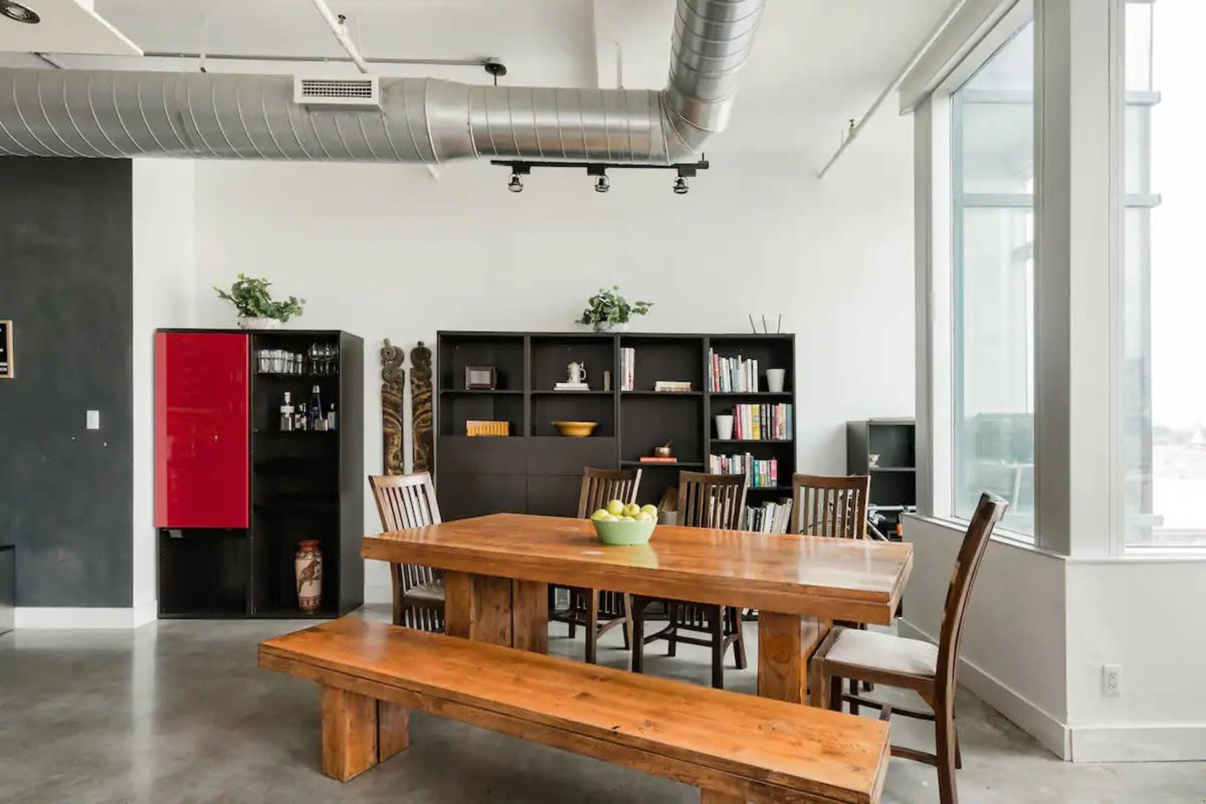 A modern dining area with a large wooden table and a row of chairs, flanked by a bookshelf and a red refrigerator against a gray wall.