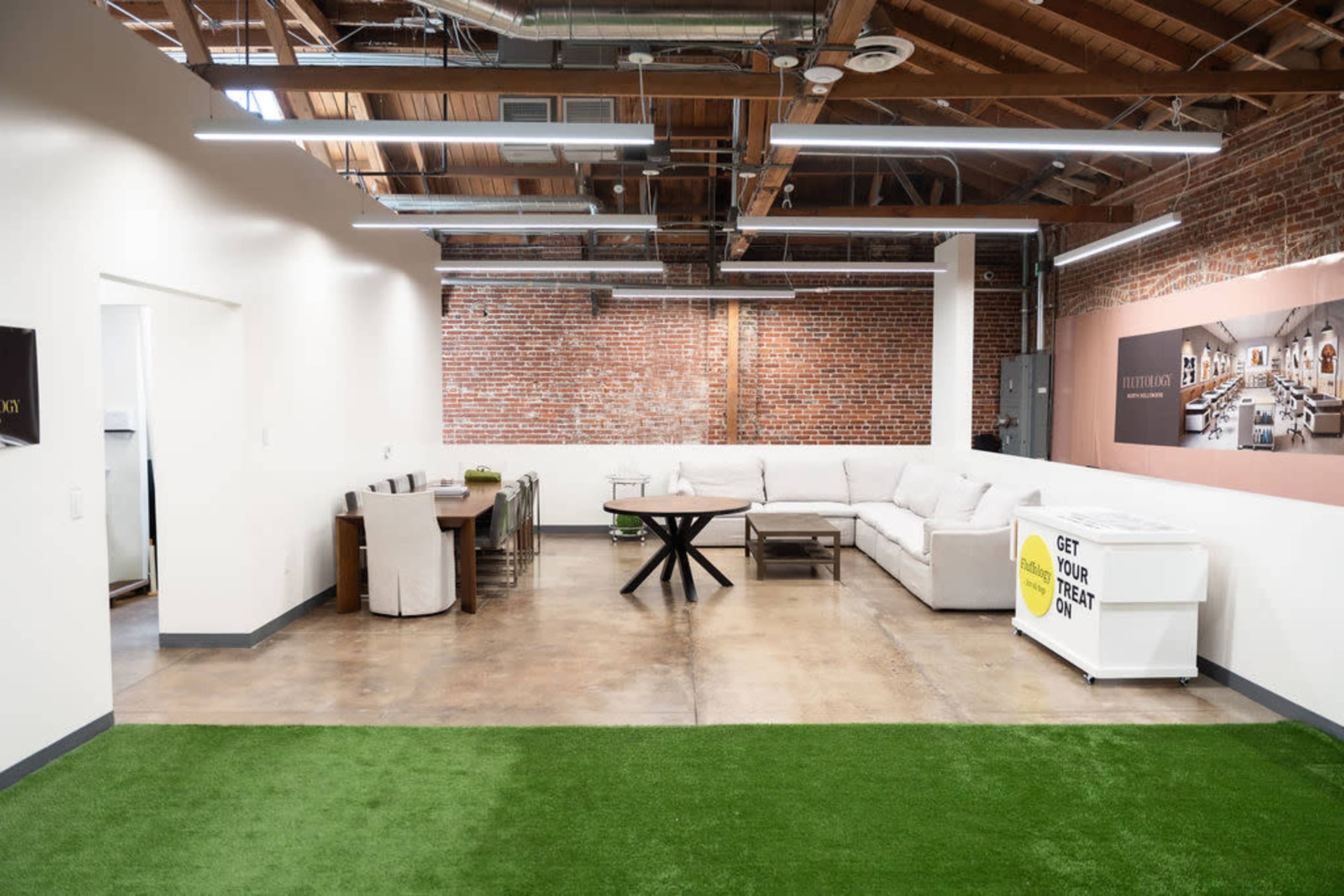 The image shows an open office space featuring a seating area with a large sectional sofa, a dining table, and sections of artificial grass on the floor.